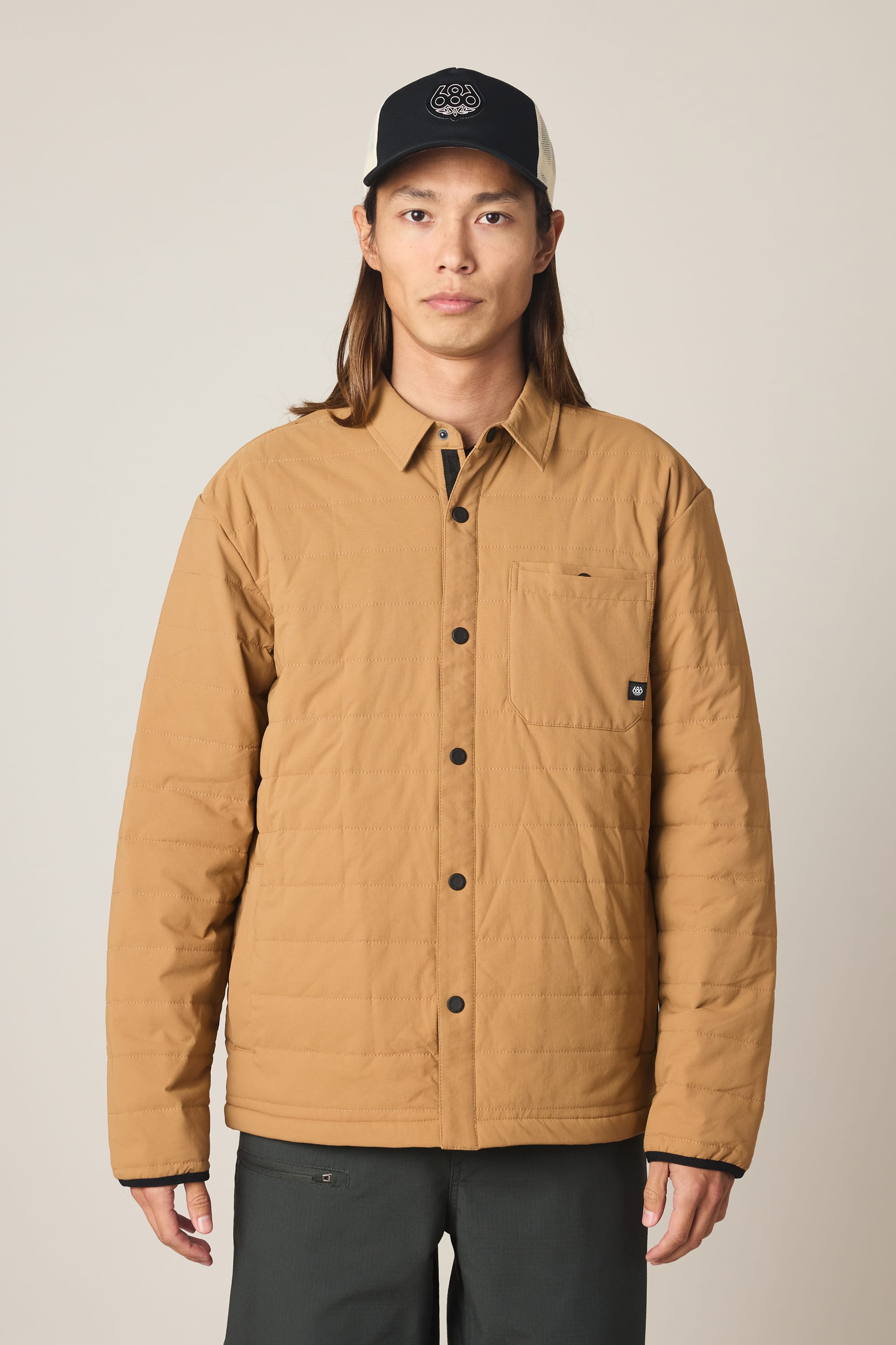 A person with long brown hair wears a tan 686 Men's Thermaldry Insulated Shacket, dark pants, and a black-and-white cap, standing against a plain light background.