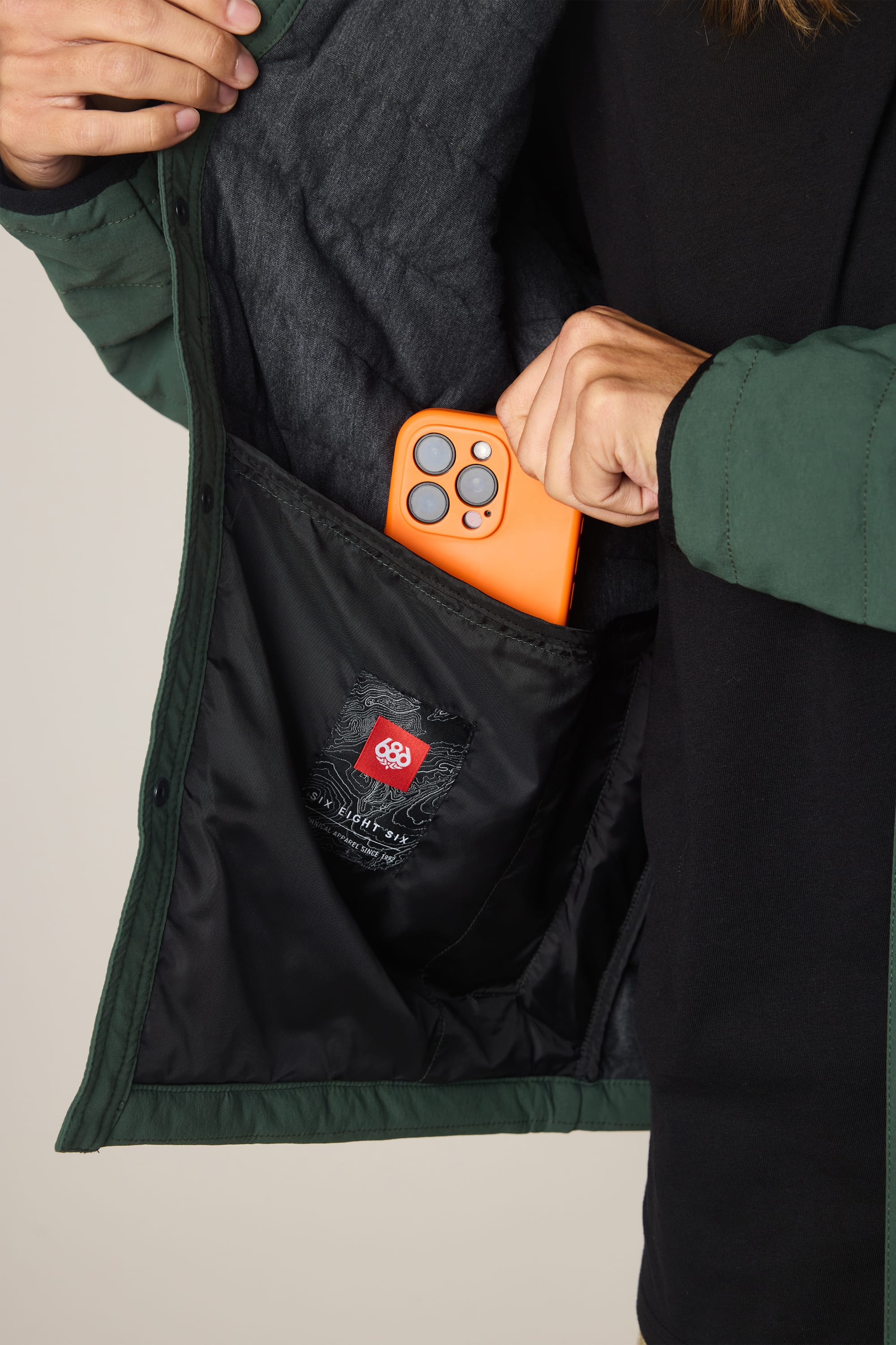 A person slips an orange smartphone into the inner pocket of a partially open 686 Men's Thermaldry Insulated Shacket, revealing the phone and the pocket’s fabric label.