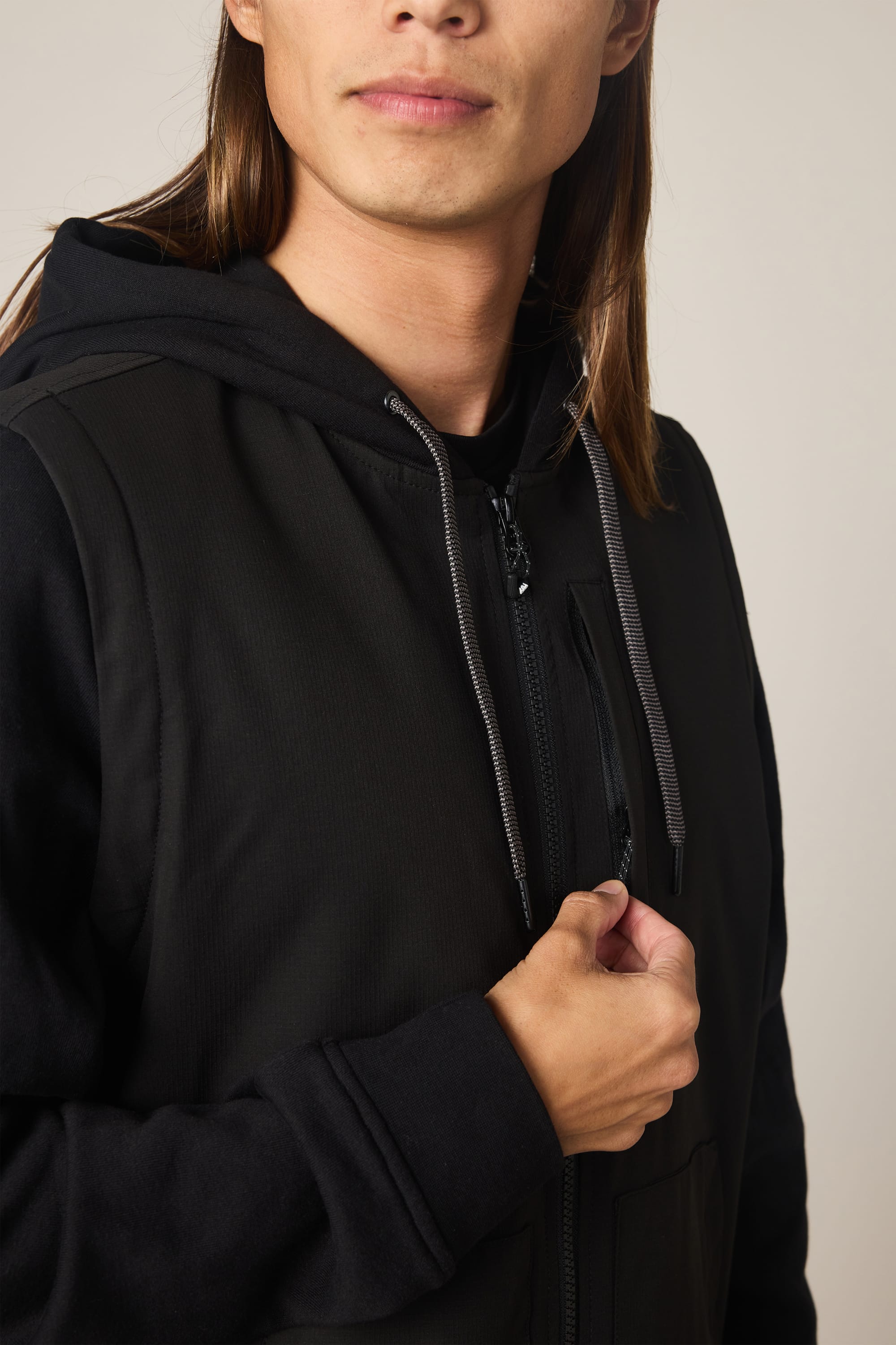 A person with long brown hair, shown from the shoulders up against a plain, neutral background, is wearing the 686 Men's Merge Quilted Hoody by 686 and holding the zipper near their chest.
