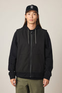 A person with long hair wears a black 686 Men's Merge Quilted Hoody, olive green pants, and a black baseball cap with a white logo, standing against a plain light background.