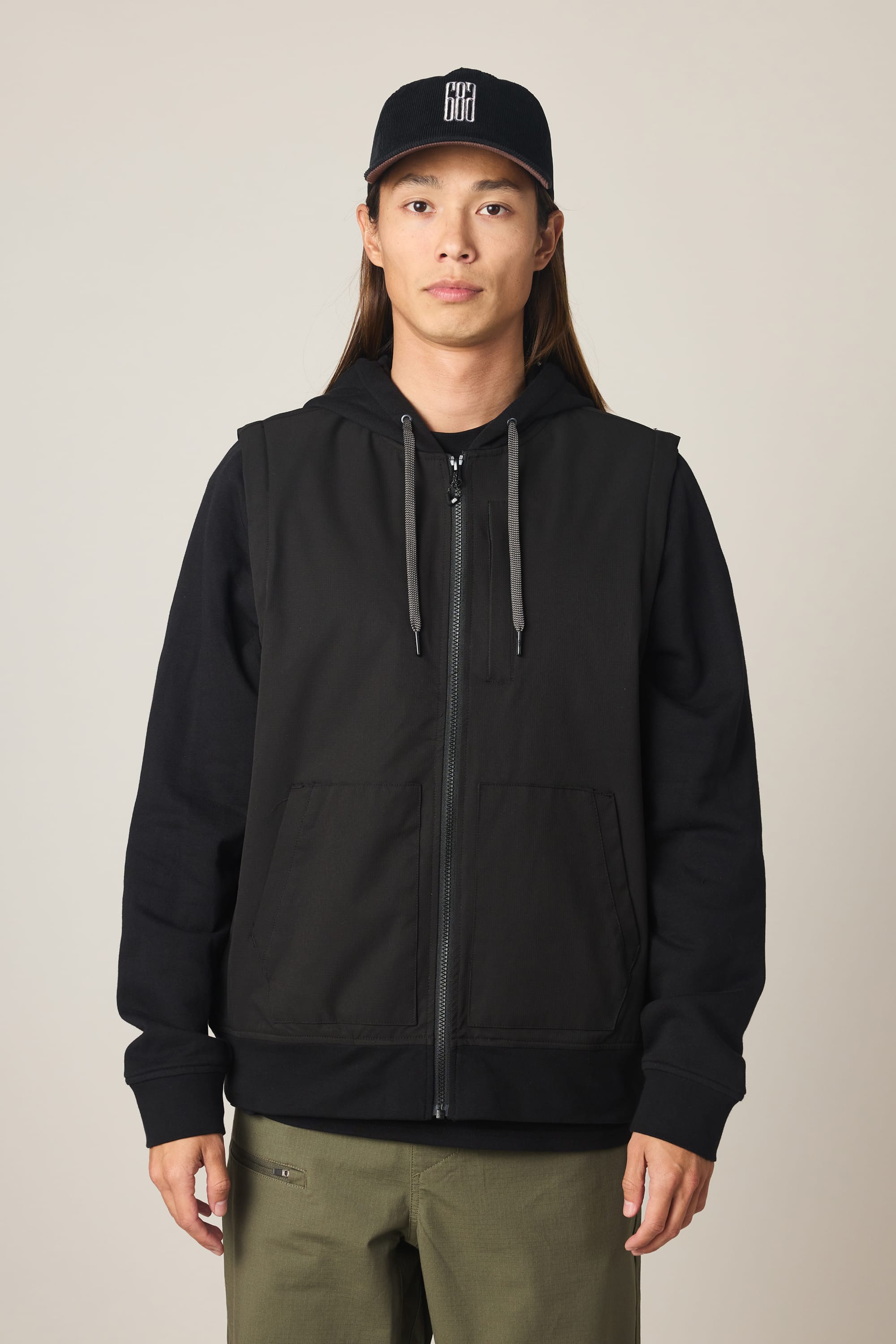 A person with long hair wears a black 686 Men's Merge Quilted Hoody, olive green pants, and a black baseball cap with a white logo, standing against a plain light background.