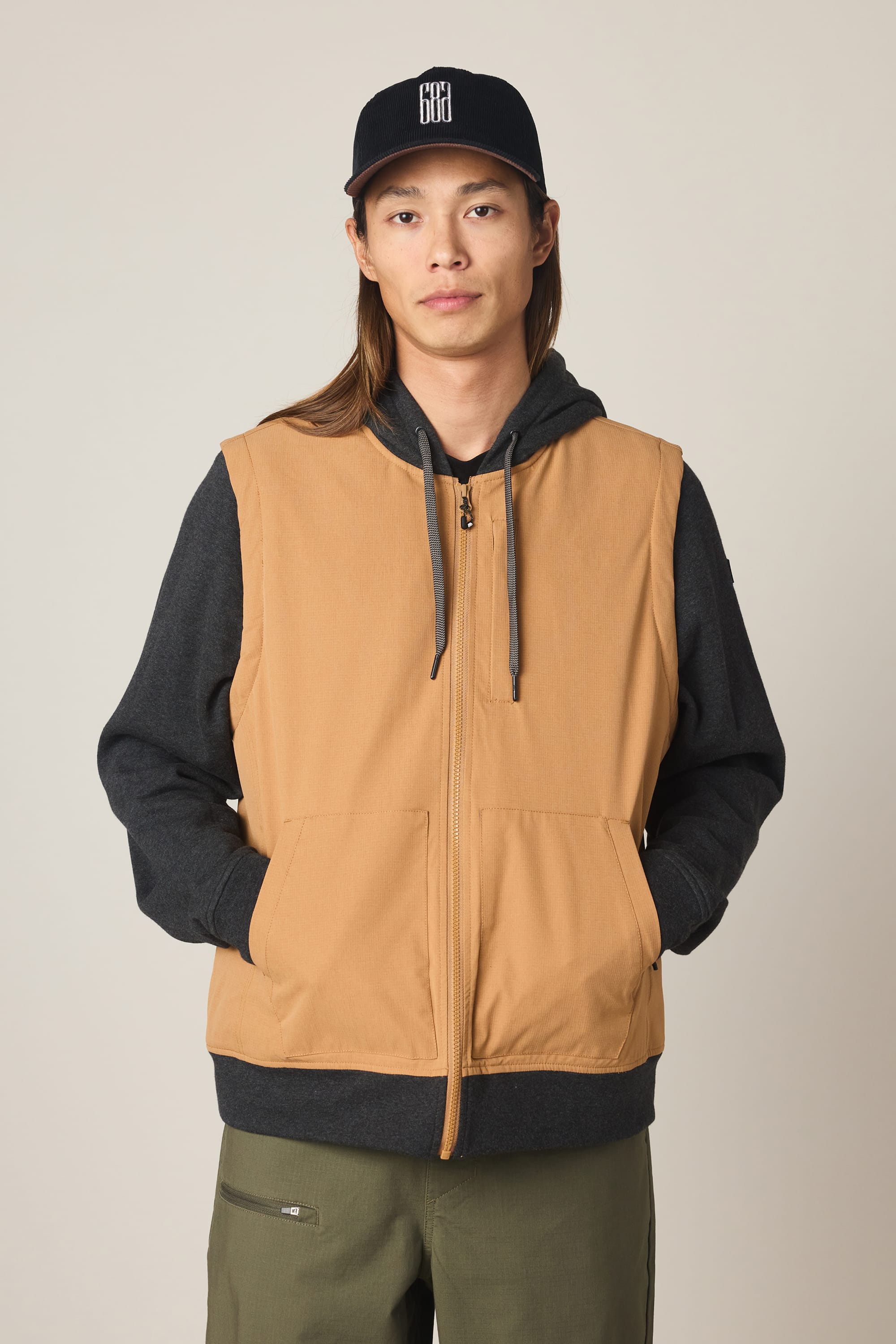 A person wears the 686 Men's Merge Quilted Hoody under a tan jacket, paired with olive green pants and a black cap, standing with hands in vest pockets against a plain light background.