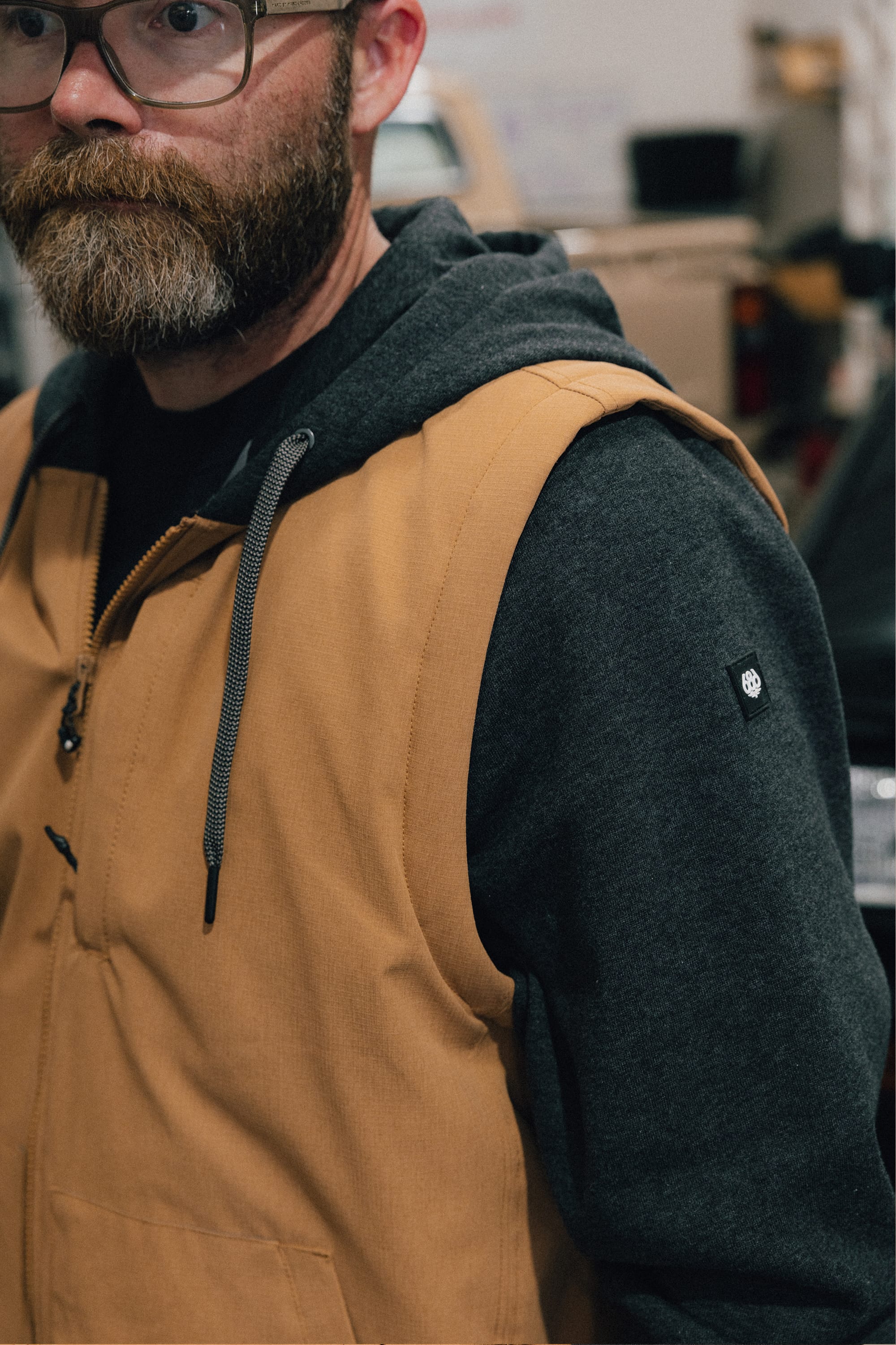 A bearded person wearing glasses, a gray hoodie, and a tan sleeveless vest stands indoors, partially facing the camera. Layered under the vest is the 686 Men's Merge Quilted Hoody from 686, adding both style and comfort to his look.