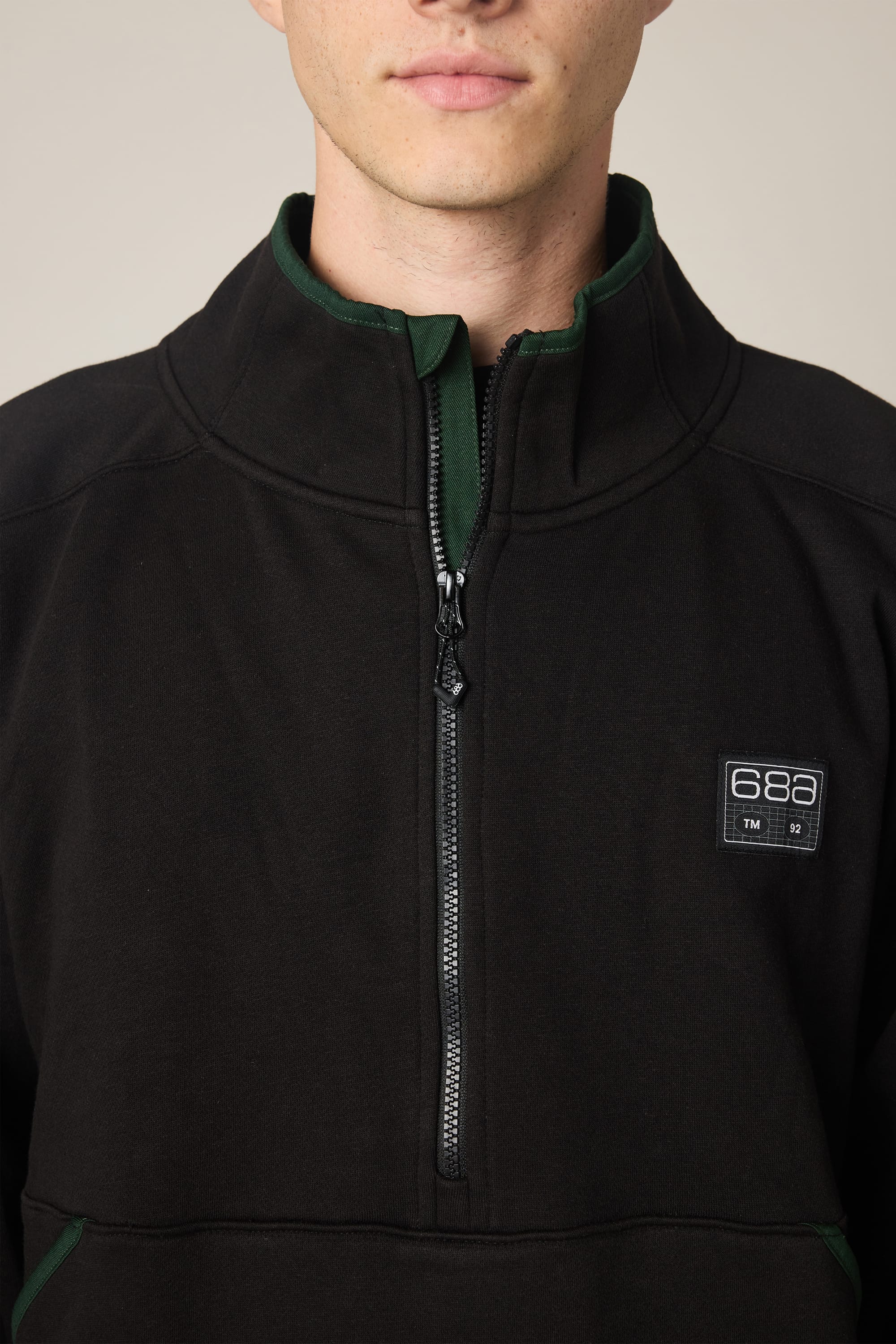 A person wearing the 686 Men’s Burnout Anorak—a black woven jacket with green trim and a 686 logo patch on the left chest. Only their lower face and upper torso are visible, set against a neutral background.