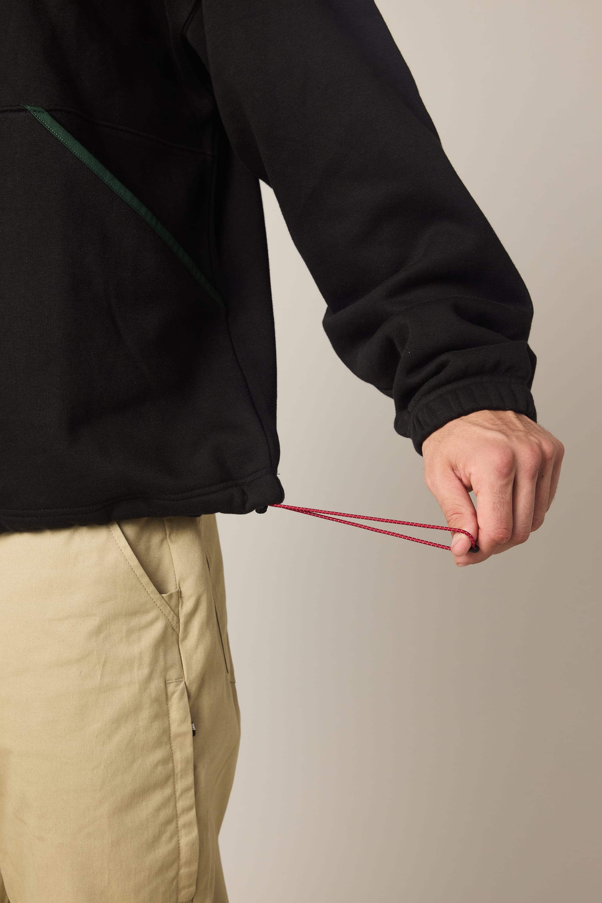 A person wearing the 686 Men's Burnout Anorak in black and beige pants adjusts the red drawstring at the jacket's hem, tightening it with one hand.