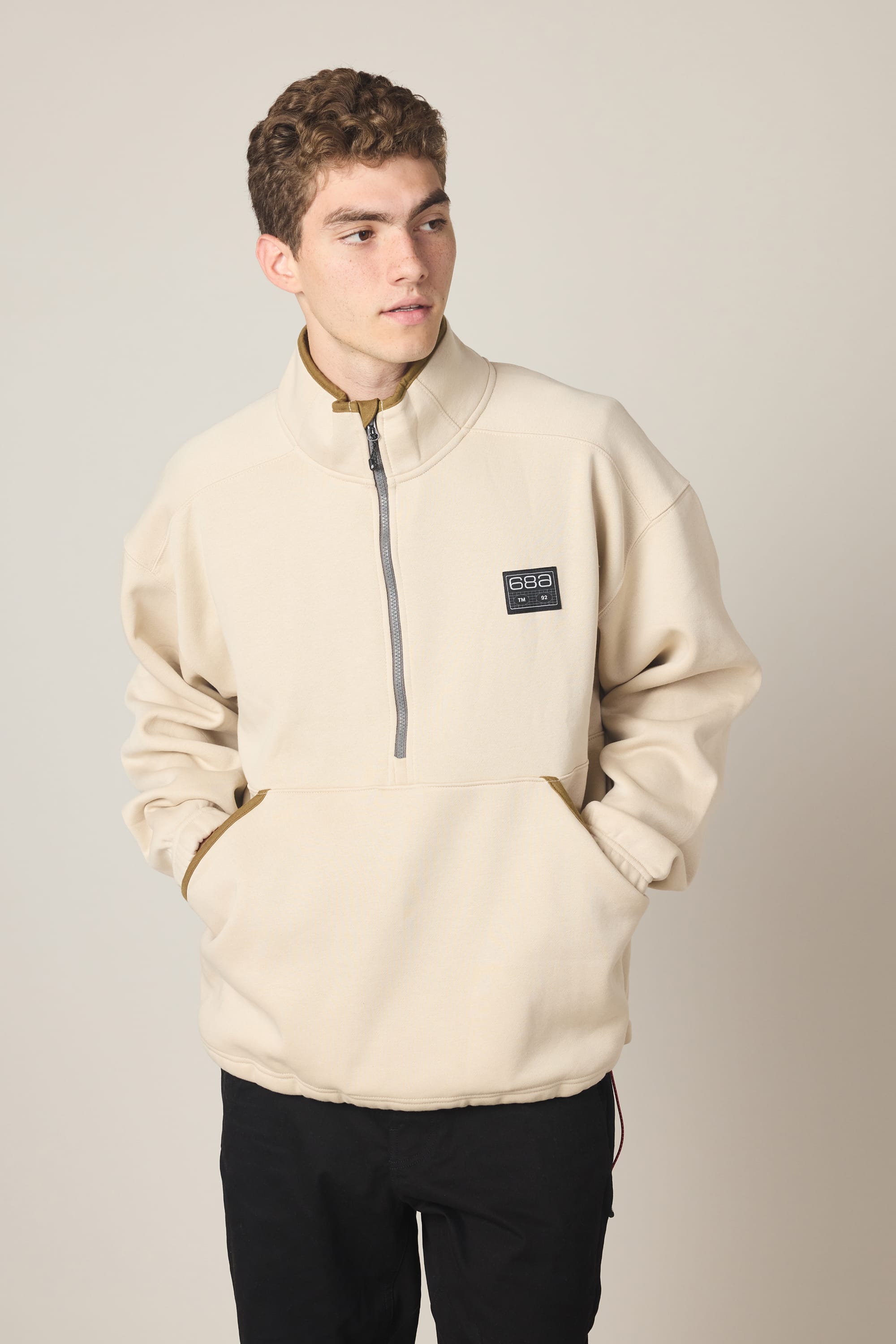 A young man stands with hands in pockets, wearing the 686 Men's Burnout Anorak—a beige woven half-zip jacket with a black chest patch—looking to the side against a plain light background.