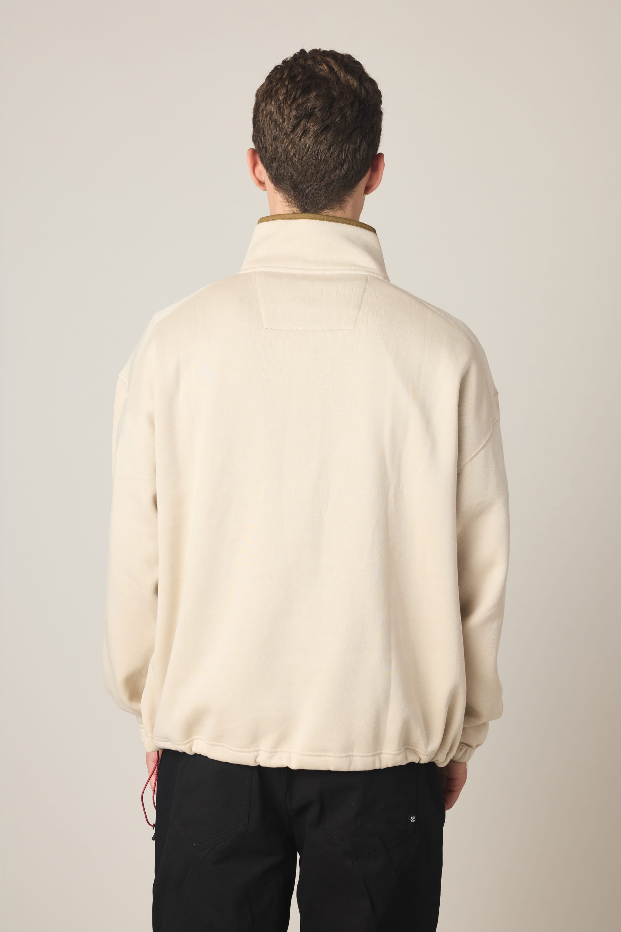A person with short brown hair faces away, wearing the 686 Men's Burnout Anorak by 686 and black pants against a plain, light background.
