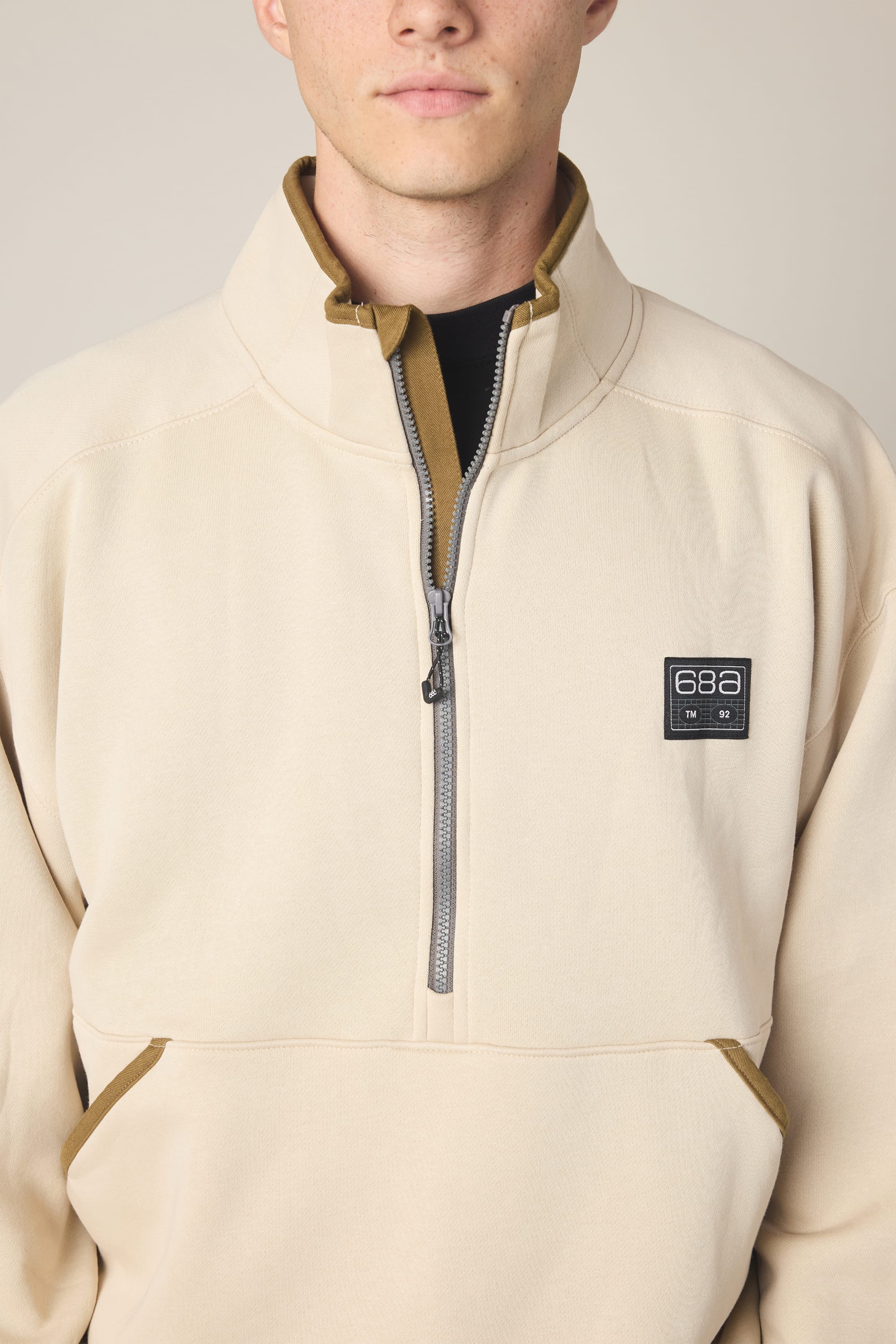 A person wearing the 686 Men's Burnout Anorak—a beige zip-up sweatshirt with a high collar, brown accents, and a 686 logo patch on the left chest, styled to resemble a men's woven jacket and crafted from polyester waterproof jacket material—standing against a neutral background.
