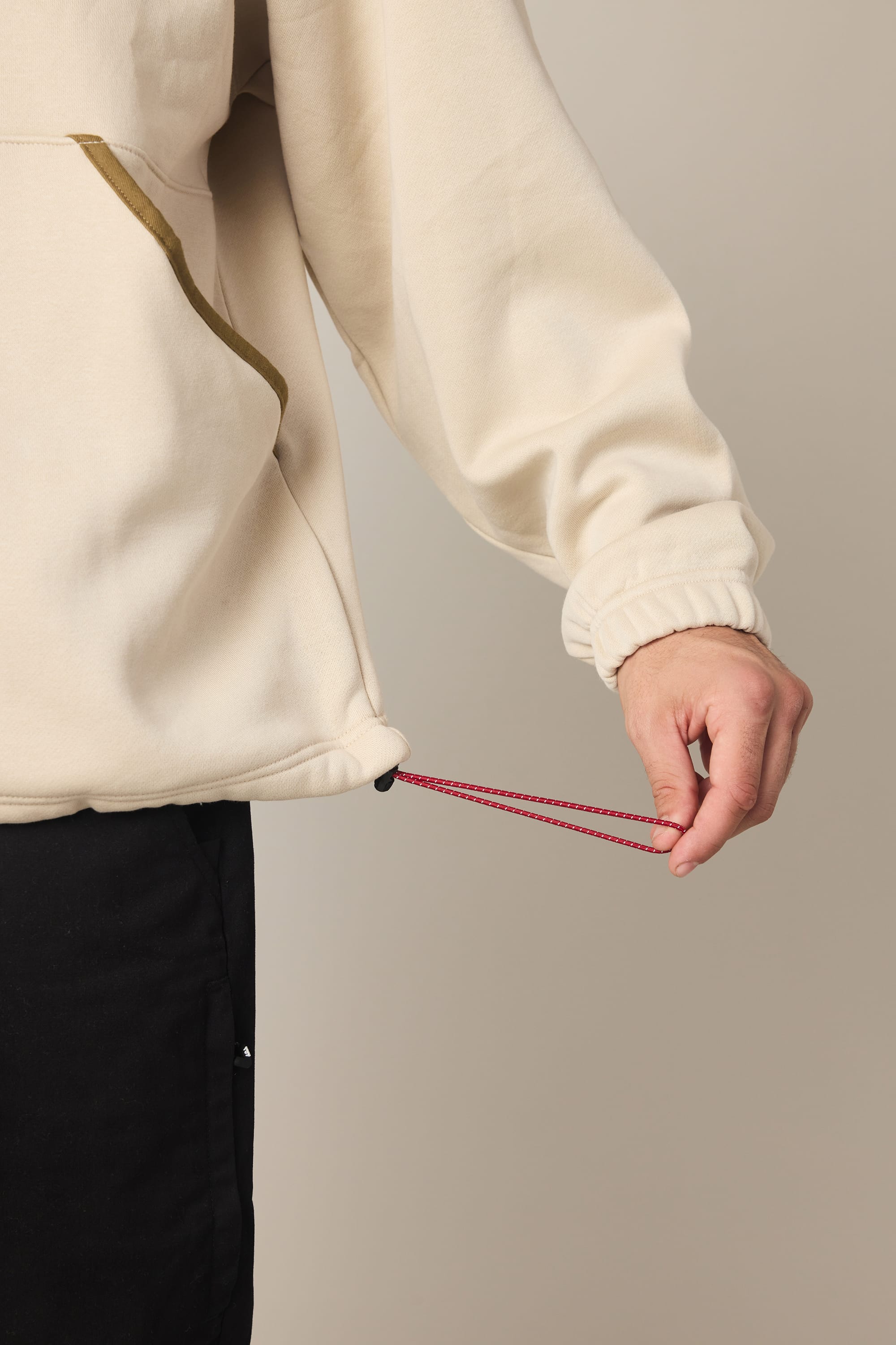 A person tightens the red drawstring at the hem of a beige 686 Men's Burnout Anorak, showing only their torso and one hand, paired with black pants.