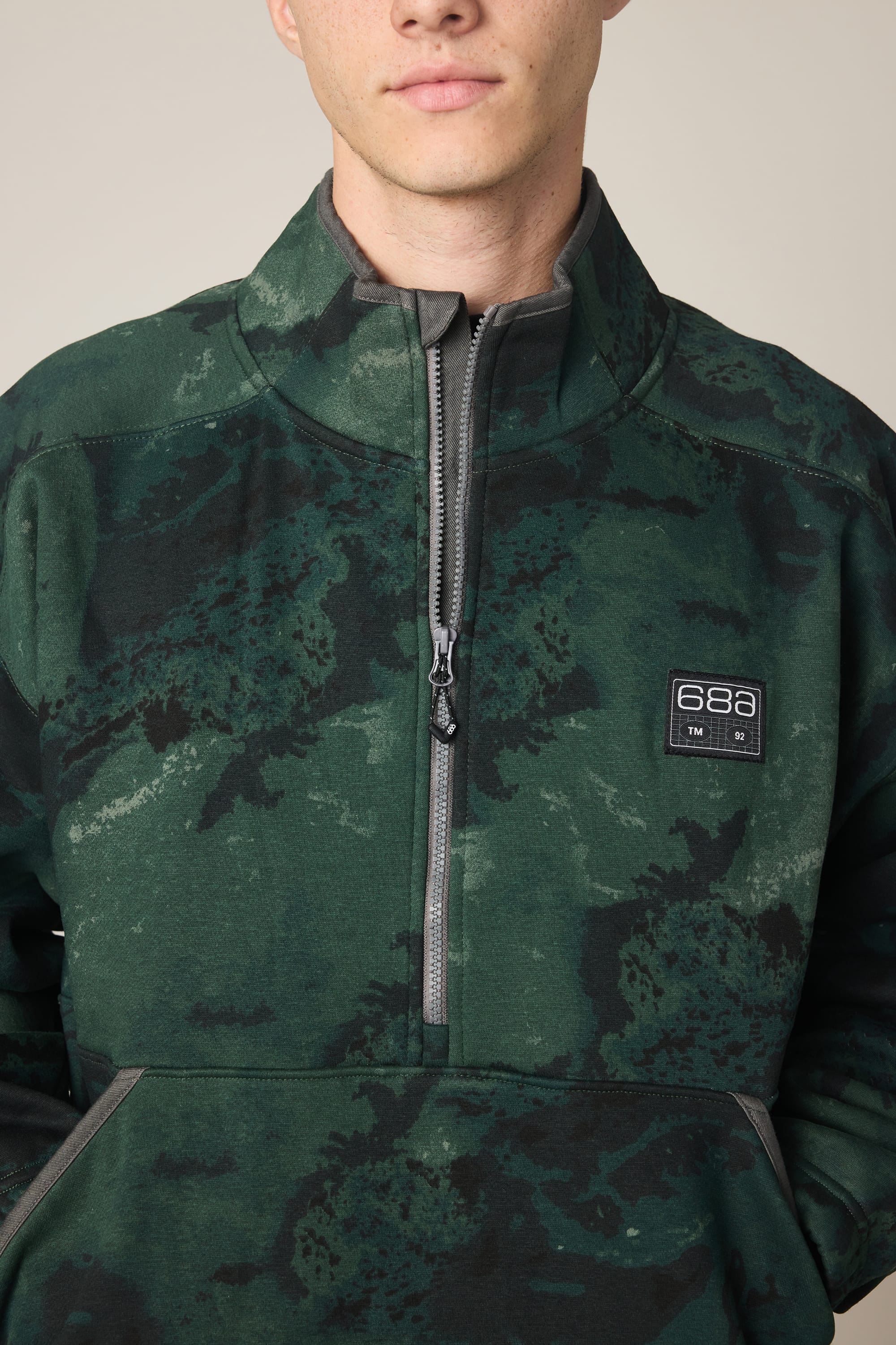 A person wearing the 686 Men's Burnout Anorak—a dark green camo-patterned polyester jacket with a half-zip front and 686 patch on the left chest. Only their lower face and upper torso are visible.