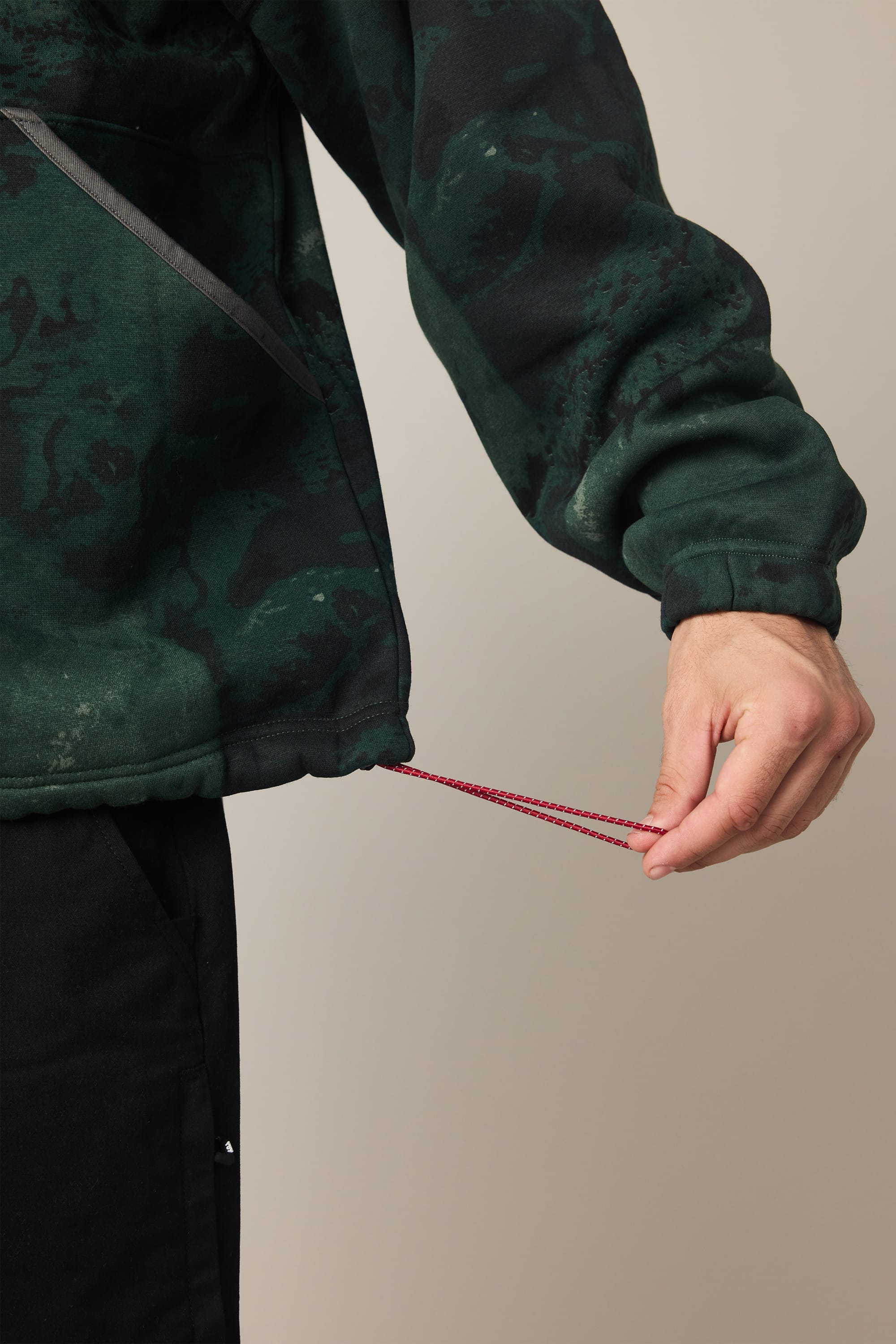 A person adjusts the red drawstring at the hem of a 686 Men's Burnout Anorak by 686, featuring a green and black camouflage pattern; only their hand and part of their body are visible.