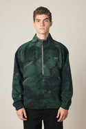 A young man with short brown hair wears the 686 Men's Burnout Anorak, a dark green camouflage half-zip jacket with a small 686 logo on the chest, standing against a plain light background.