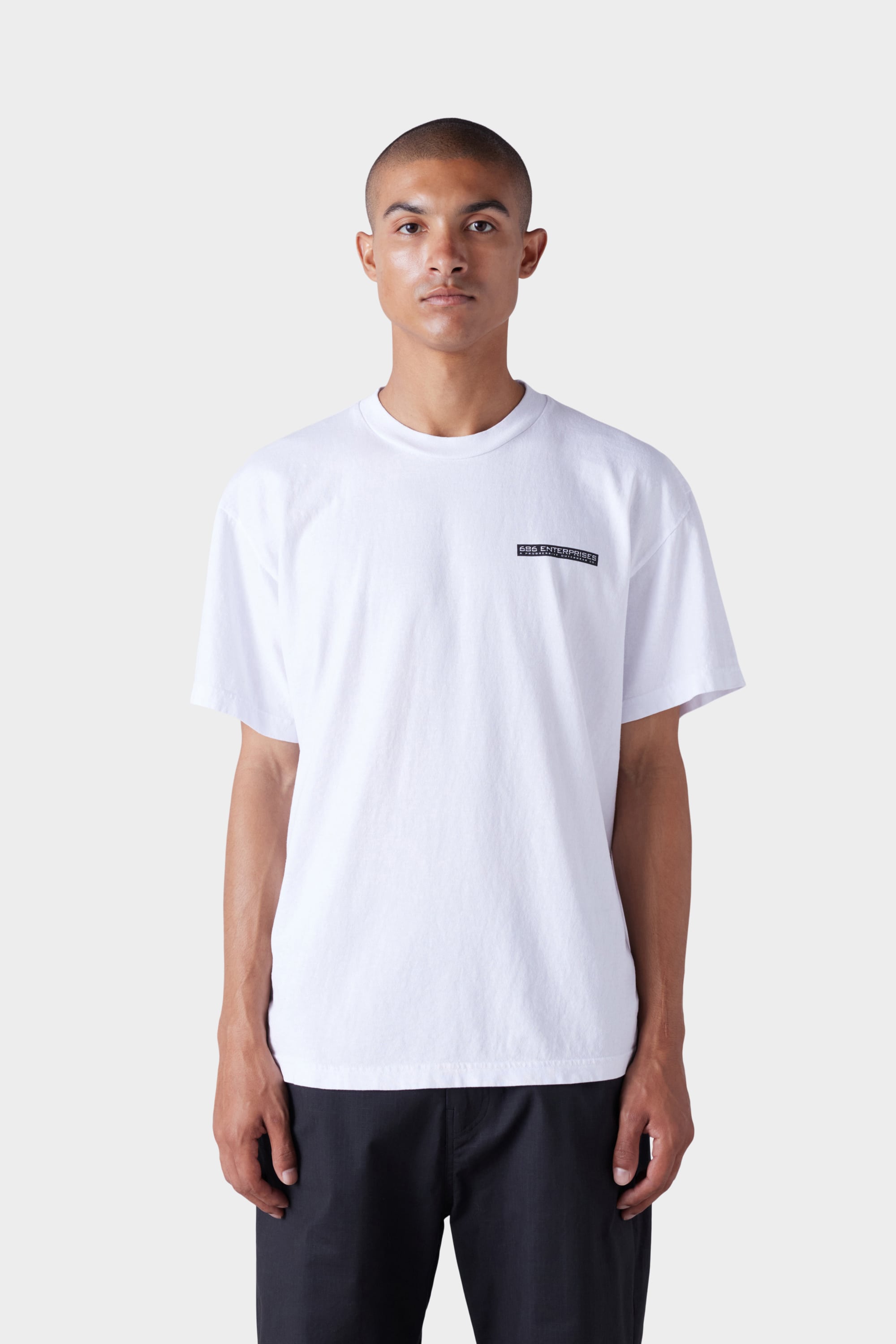 A person wears the 686 Mens Global Enterprises Short Sleeve Tee in white with small black text on the left chest and black pants, standing facing forward against a light gray background.