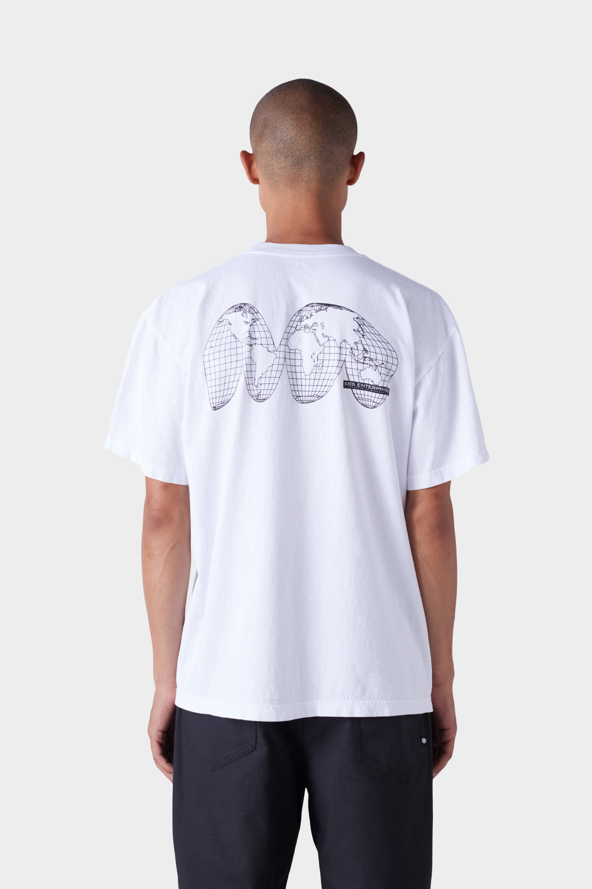 A person with short hair wears the 686 Mens Global Enterprises Short Sleeve Tee, featuring a black abstract world map graphic on the back. They pair it with vintage fit dark pants and stand against a plain, light background. Brand: 686.
