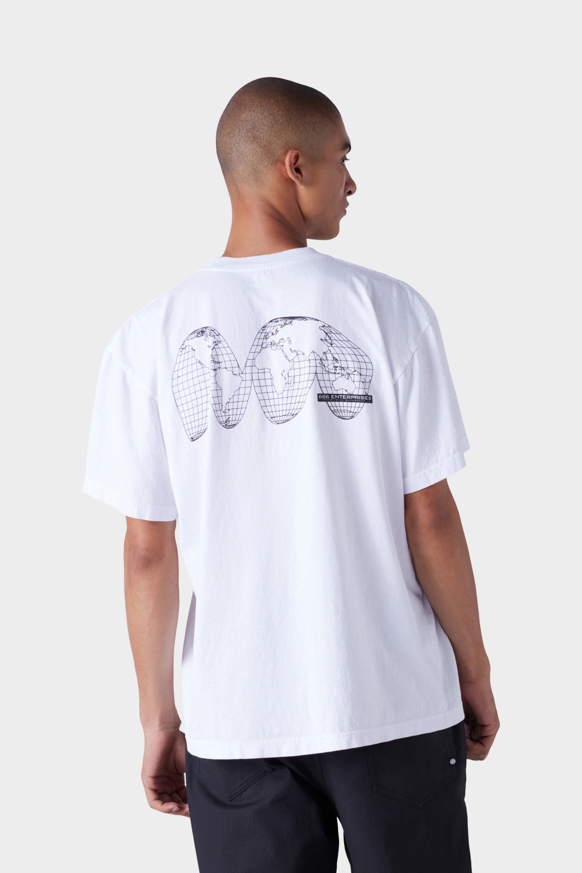 A person wearing the 686 Mens Global Enterprises Short Sleeve Tee, a loose white 686 graphic tee featuring a black abstract world map on the back, stands against a plain light background, facing away from the camera.