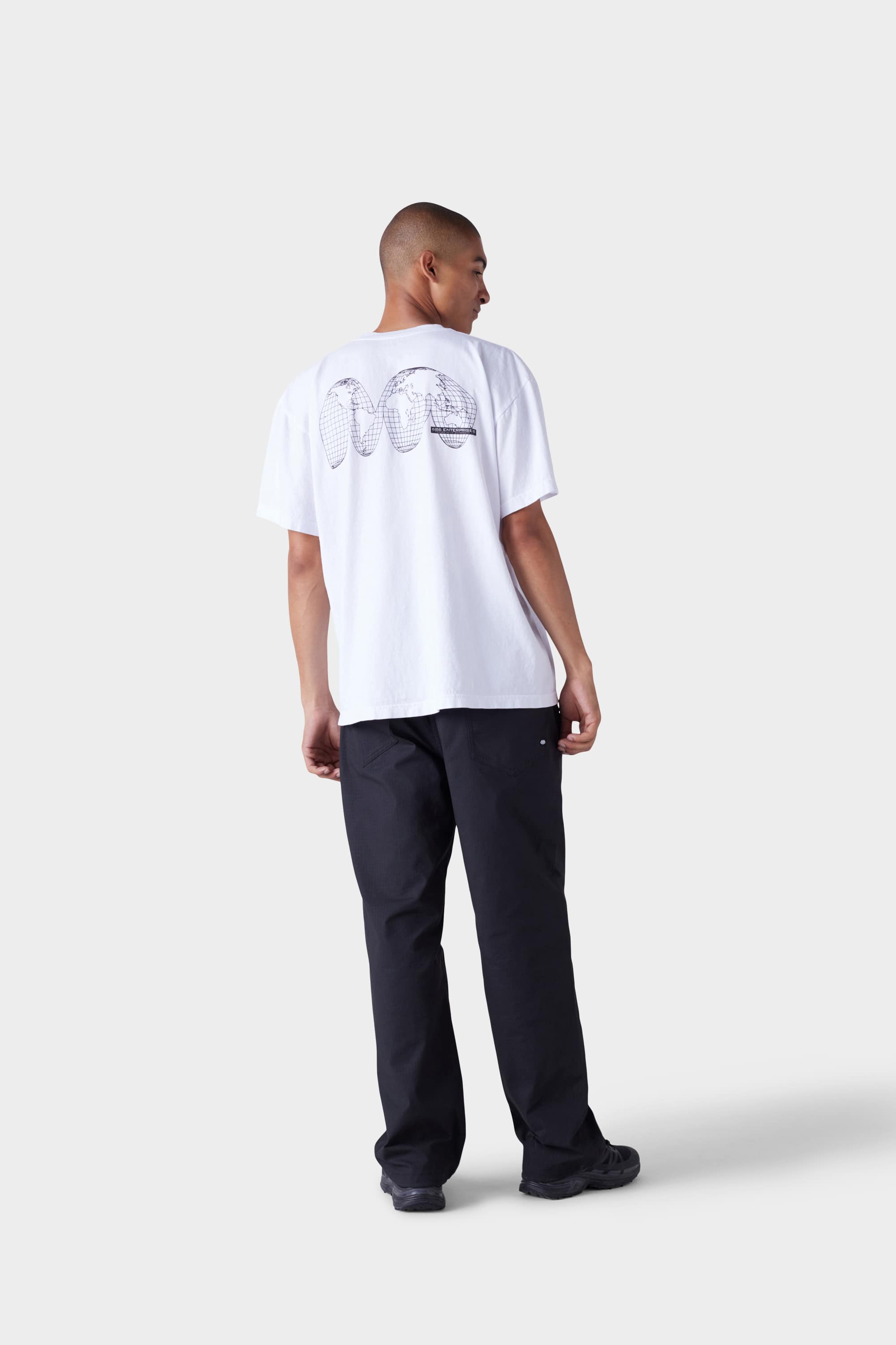 A man stands with his back to the camera, wearing the 686 Mens Global Enterprises Short Sleeve Tee by 686, featuring a black graphic on white, paired with black pants and shoes, set against a plain light gray background.