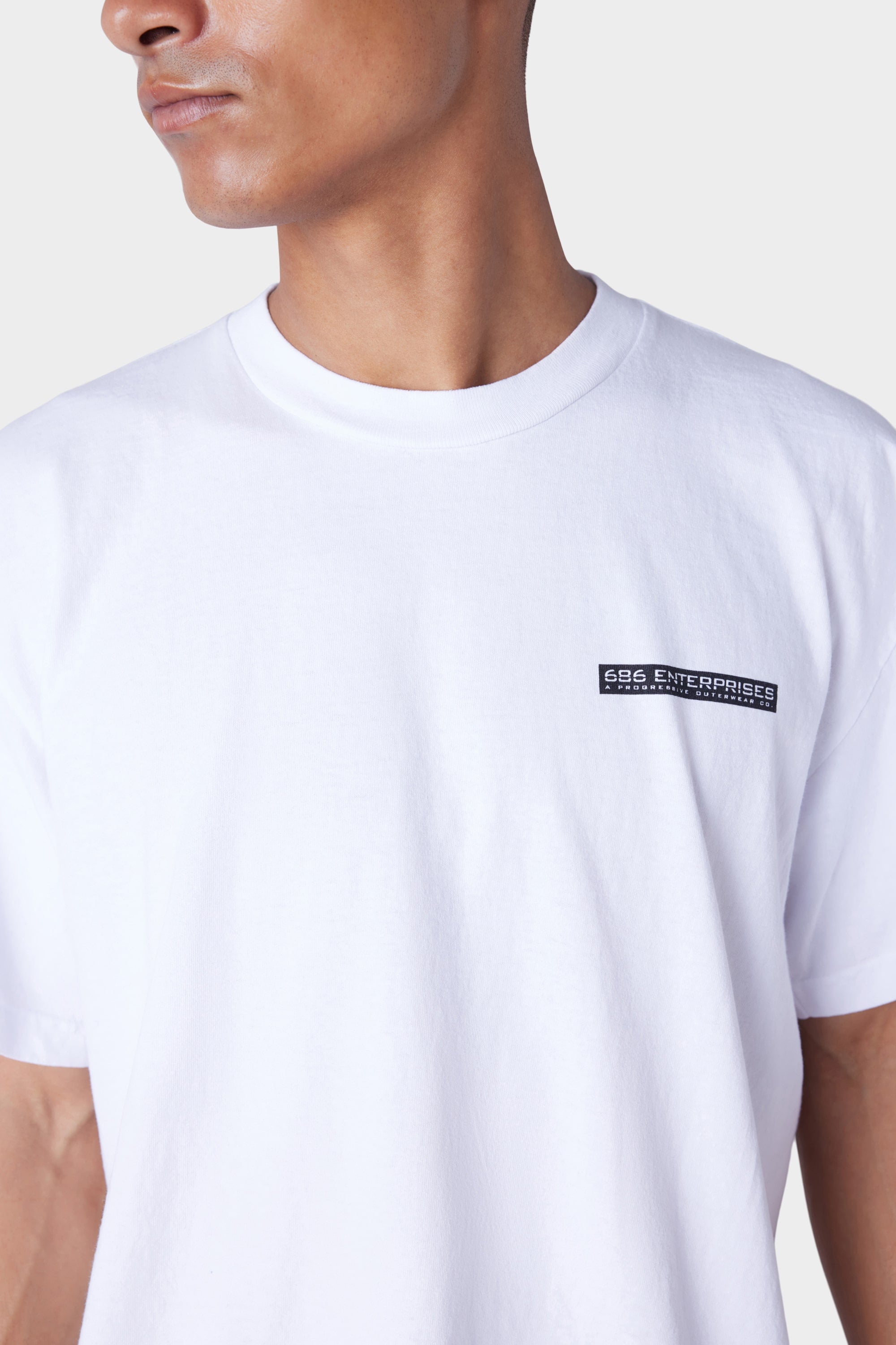 A person wears the 686 Mens Global Enterprises Short Sleeve Tee, a plain white vintage fit with a small black 686 ENTERPRISES logo on the left chest. They are turned slightly to the side, with their face partially out of frame.