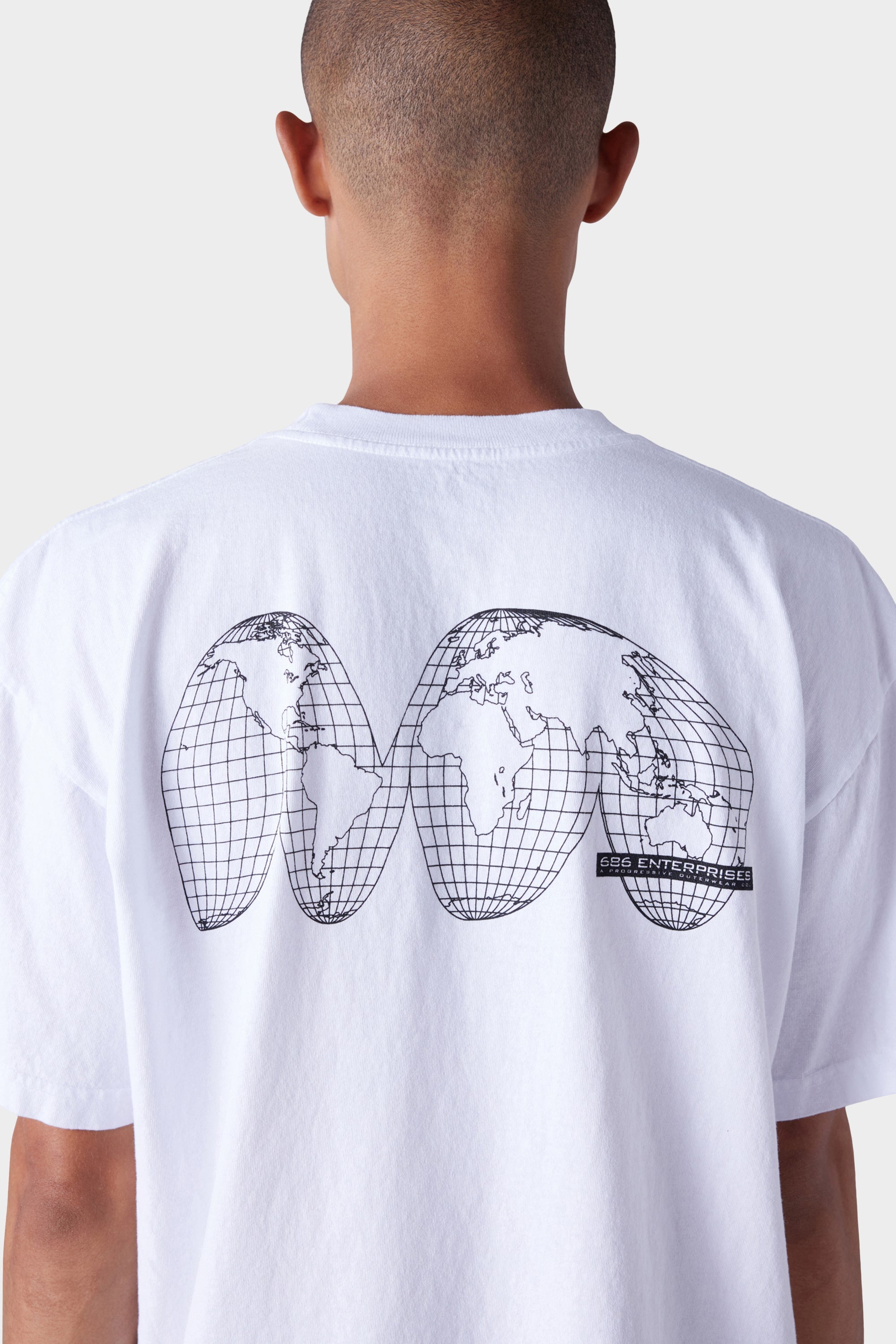 A person wears the 686 Mens Global Enterprises Short Sleeve Tee—featuring a black abstract world map and GSG ENTERPRISE text on the back—styled from behind. The vintage-fit white shirt by 686 pops against a plain, light background.
