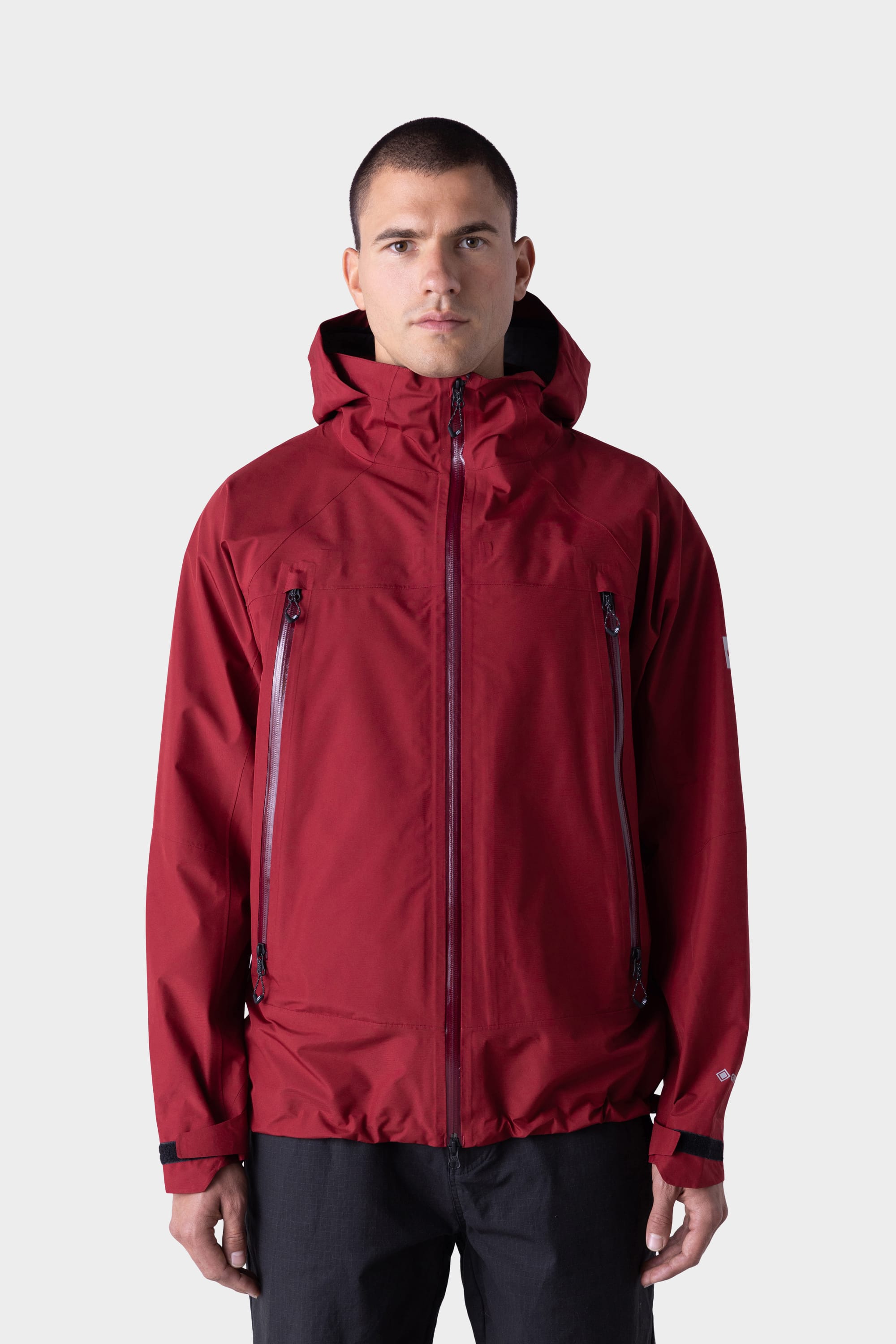 A man stands facing forward against a light gray background, wearing the 686 Mens GORE-TEX PACLITE® Jacket by 686 with black zippers. He has short hair and pairs the jacket with black pants.