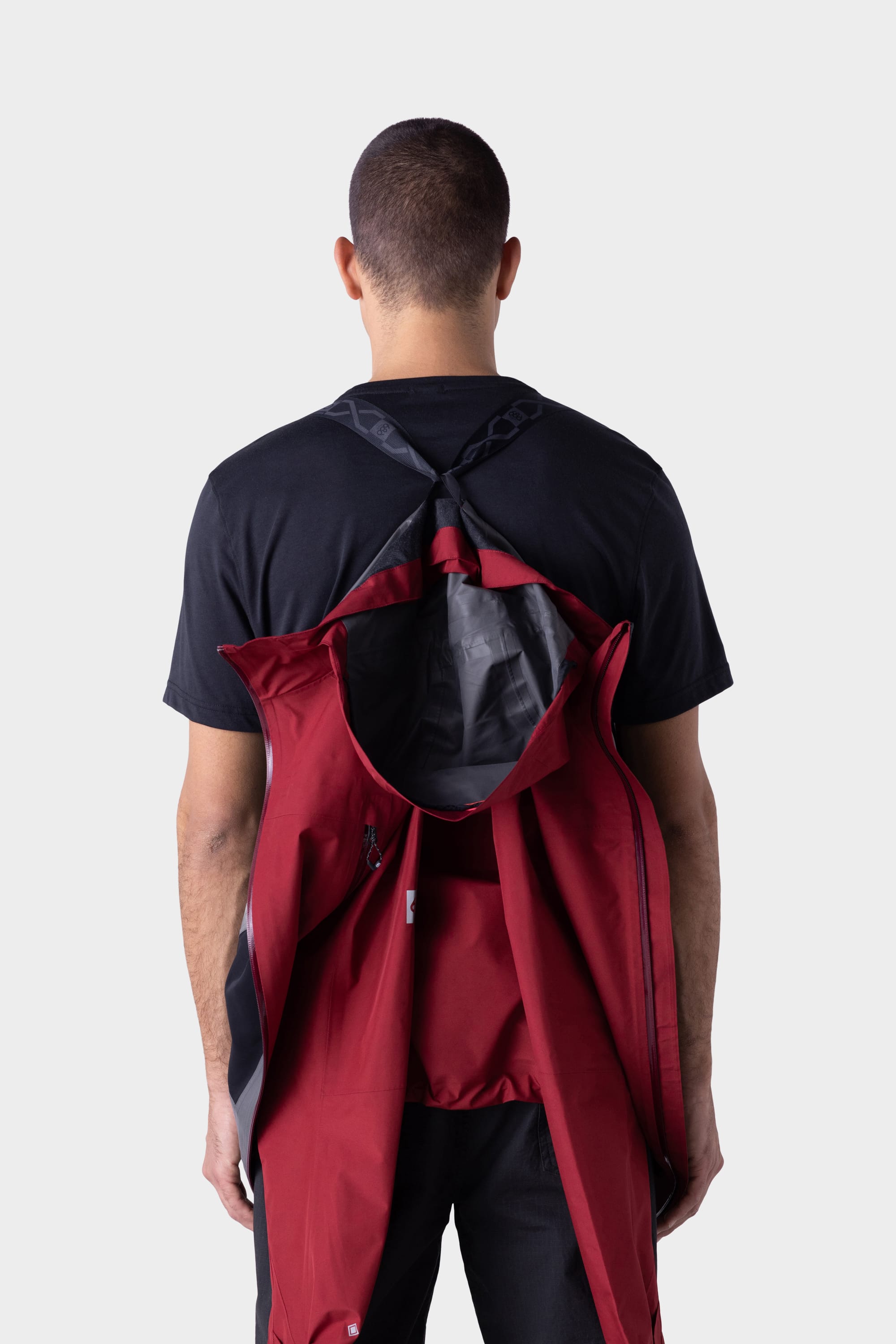 A person stands facing away in a black t-shirt and pants. The 686 Mens GORE-TEX PACLITE® Jacket, by 686, in red, is draped over their back with its collar attached to a black shoulder strap. The background is plain light gray.