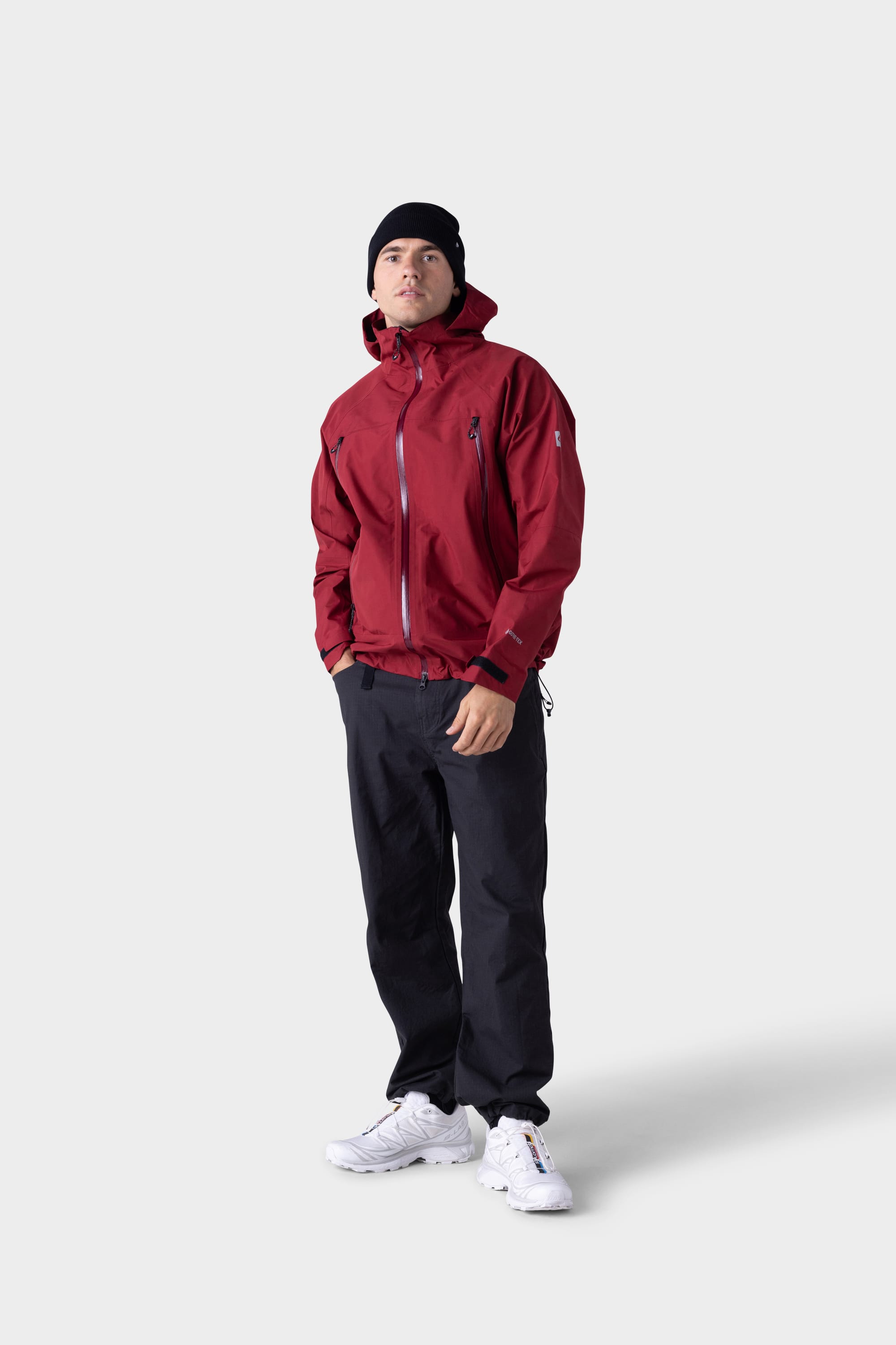 A person wearing the 686 Mens GORE-TEX PACLITE® Jacket in red, a black beanie, black pants, and white sneakers stands against a plain light gray background.