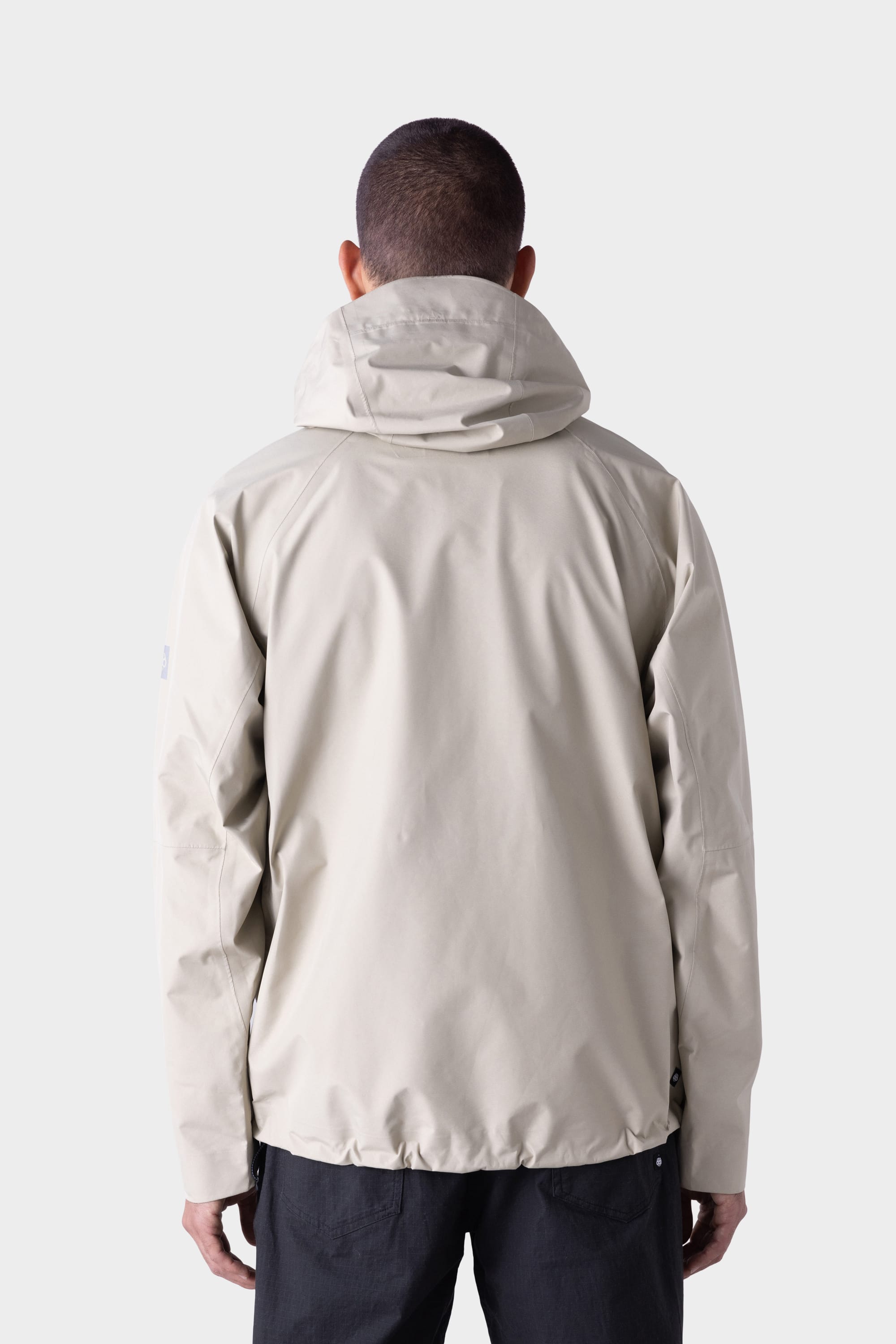 A person with short hair, seen from behind, wears the 686 Mens GORE-TEX PACLITE® Jacket in light beige and dark pants, standing against a plain, light background.