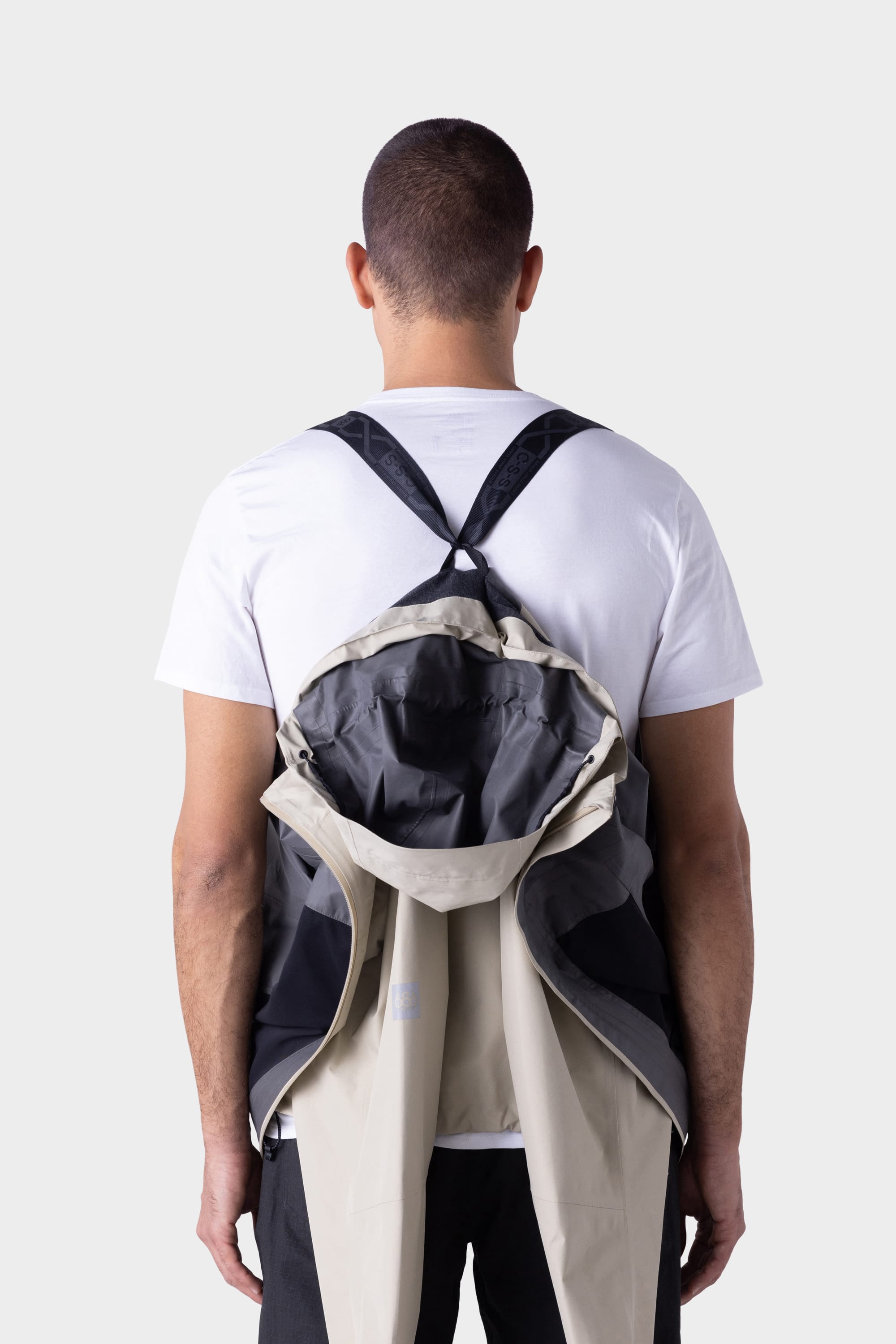 A person in a white t-shirt and black pants stands facing away. A 686 Mens GORE-TEX PACLITE® Jacket by 686, in beige and black, is draped backward over their shoulders with sleeves hanging and back open to show the interior.