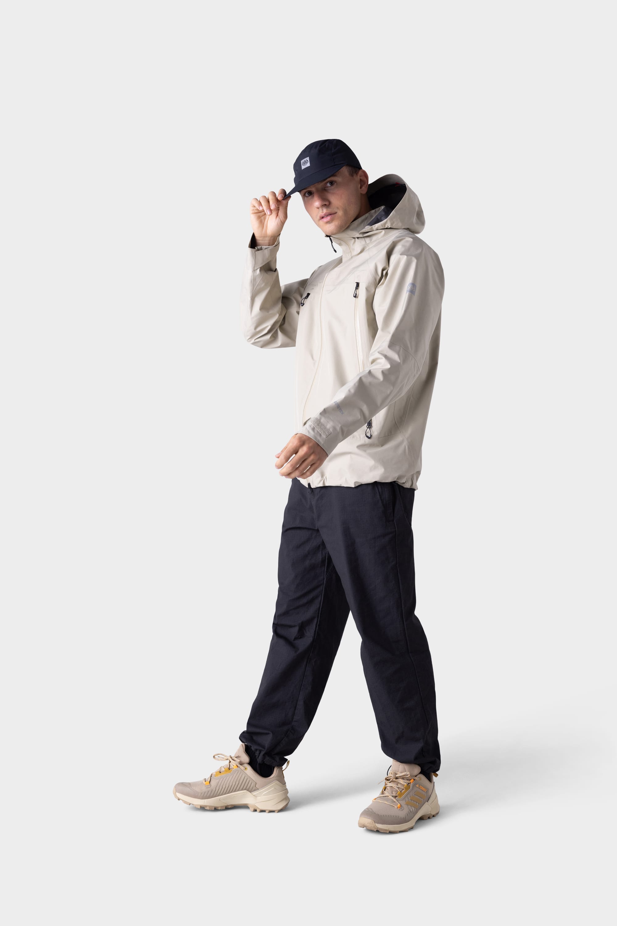 A person wearing a 686 Mens GORE-TEX PACLITE® Jacket stands against a plain white background, looking forward and adjusting a black cap with one hand.