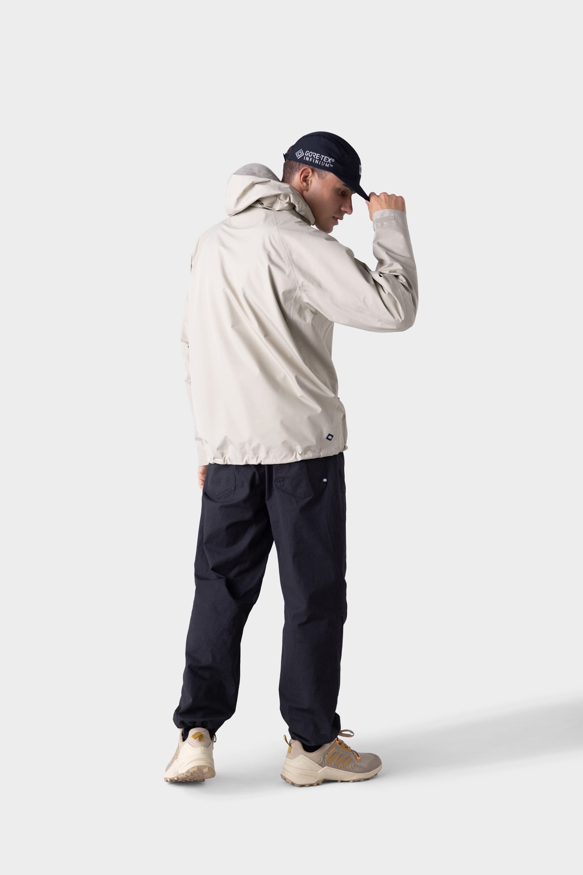 A person wearing a 686 Mens GORE-TEX PACLITE® Jacket, black pants, beige sneakers, and a black cap stands with their back to the camera, adjusting their cap against a plain white background.