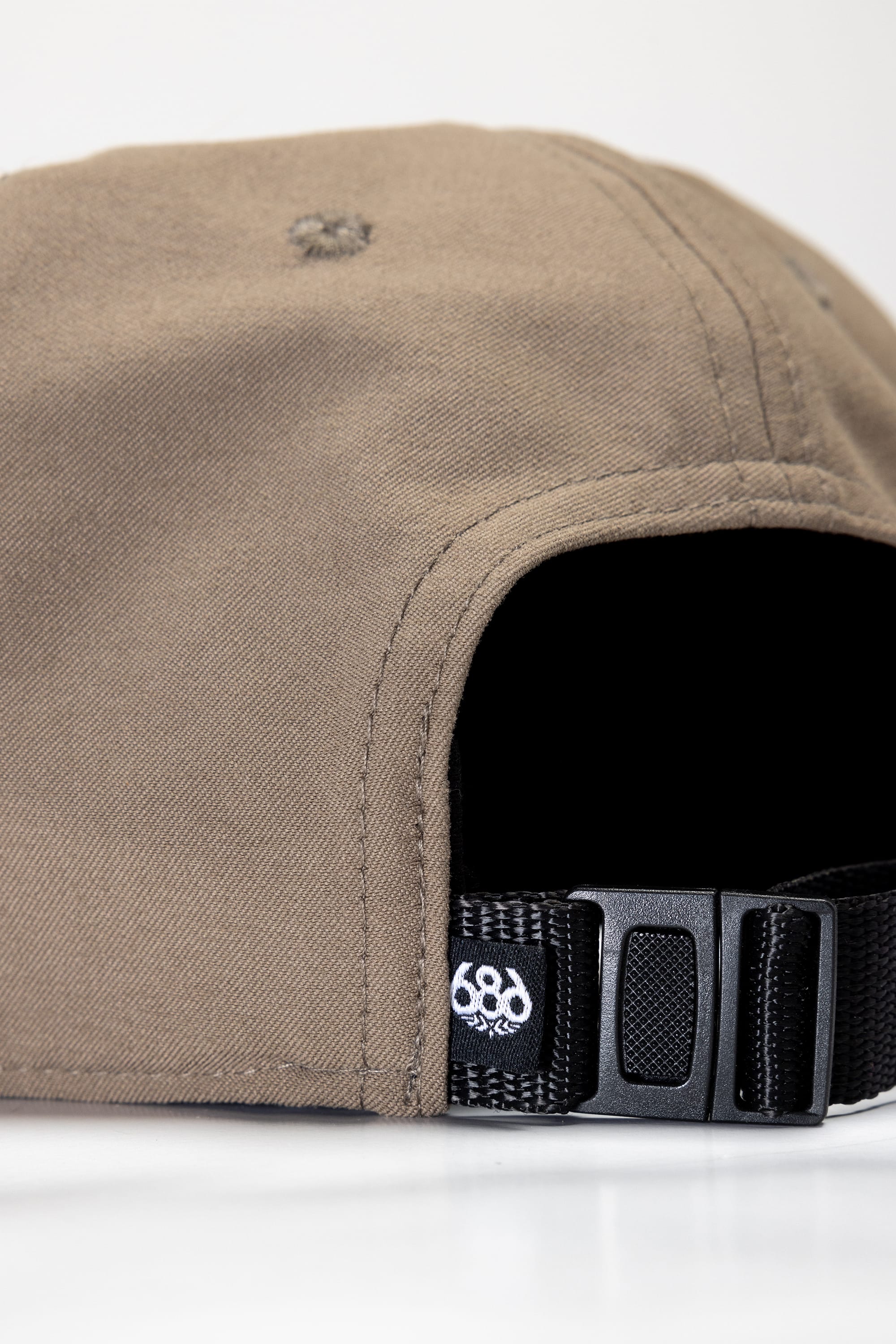 Close-up of the back of the 686 Packable Everywhere Hat by 686, showing its brown color, black adjustable strap, and a small white logo near the strap, set against a plain white background.