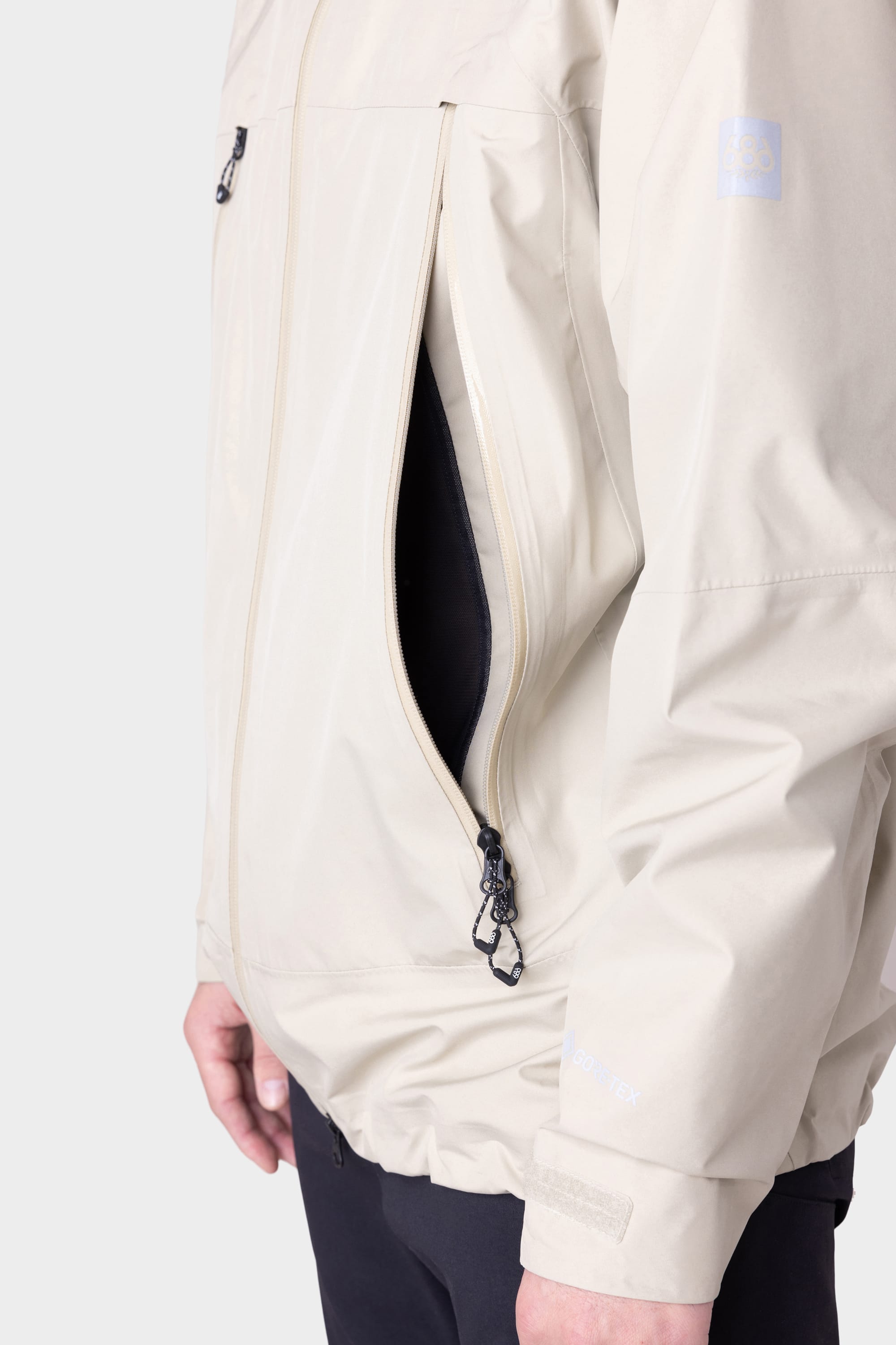 A person wears the 686 Mens GORE-TEX PACLITE® Jacket in light beige, featuring a large zippered side vent, black inner lining, and a subtle sleeve logo by 686, paired with dark pants.