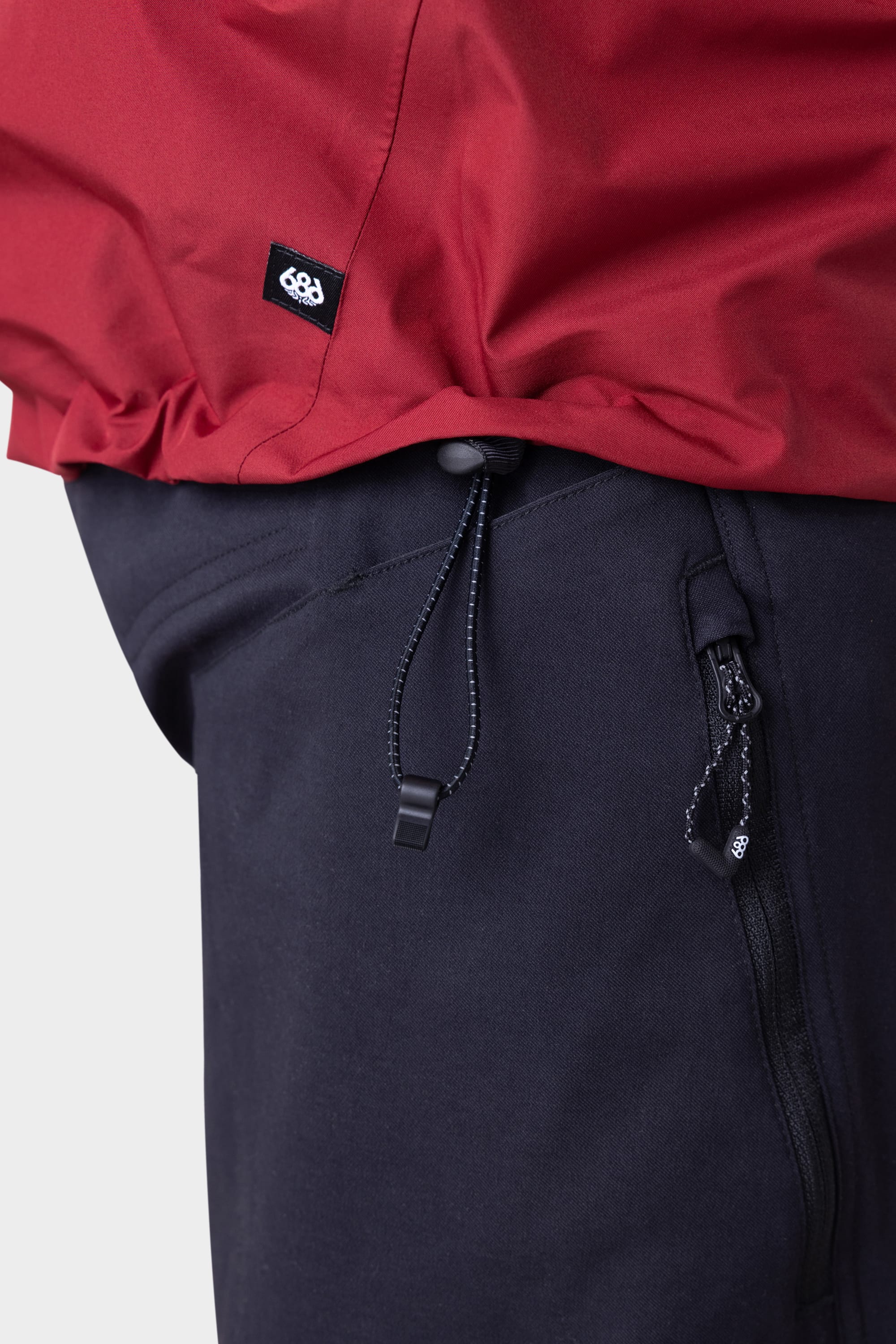Close-up of a person wearing the 686 Mens GORE-TEX PACLITE® Jacket with an adjustable drawstring hem, the ultimate versatile layer by 686 for any season, paired with dark pants featuring a zippered pocket against a plain light background.