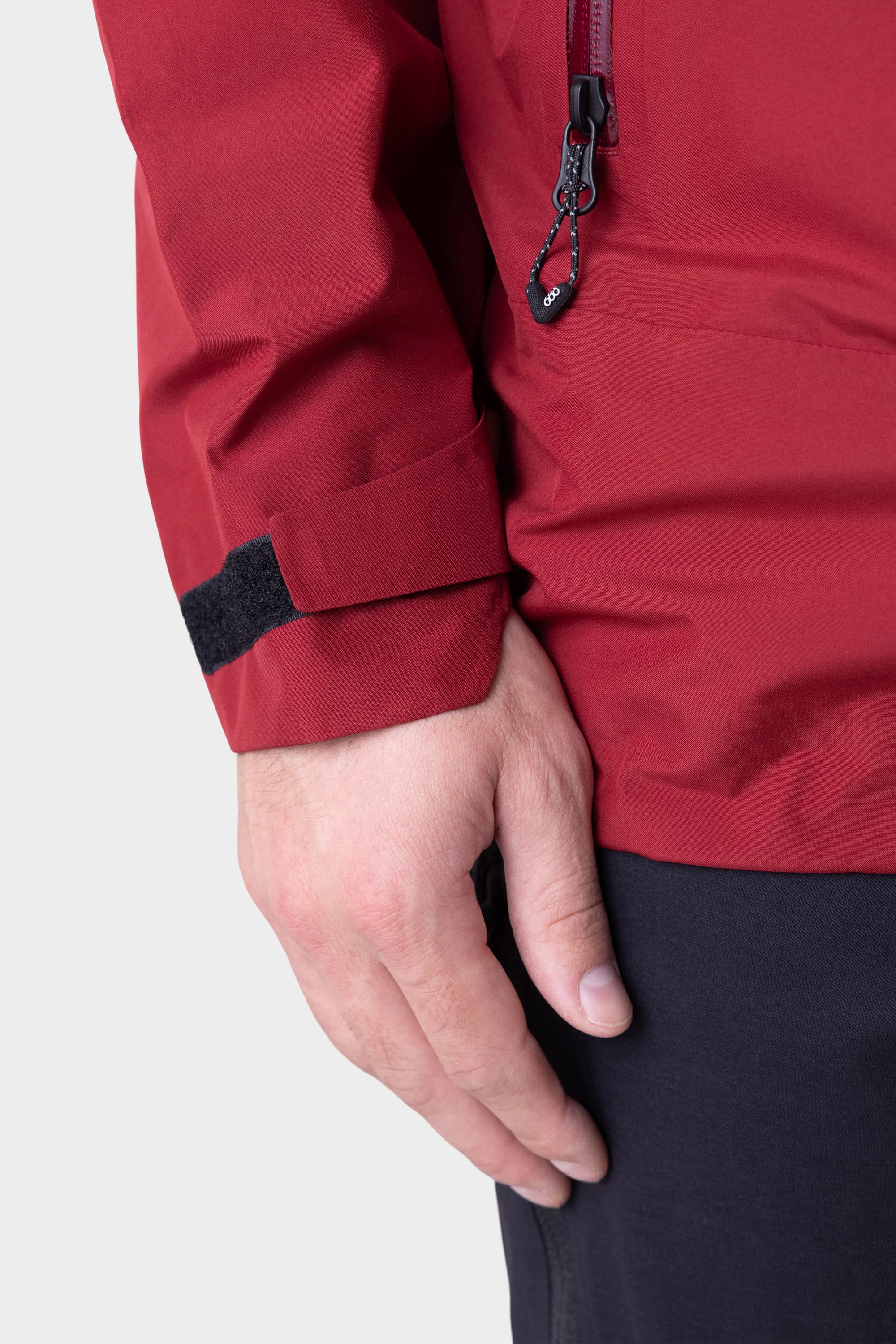A close-up of a persons left hand resting by their side, wearing the 686 Mens GORE-TEX PACLITE® Jacket with a visible zipper and black Velcro strap on the sleeve, paired with dark pants against a plain light background.