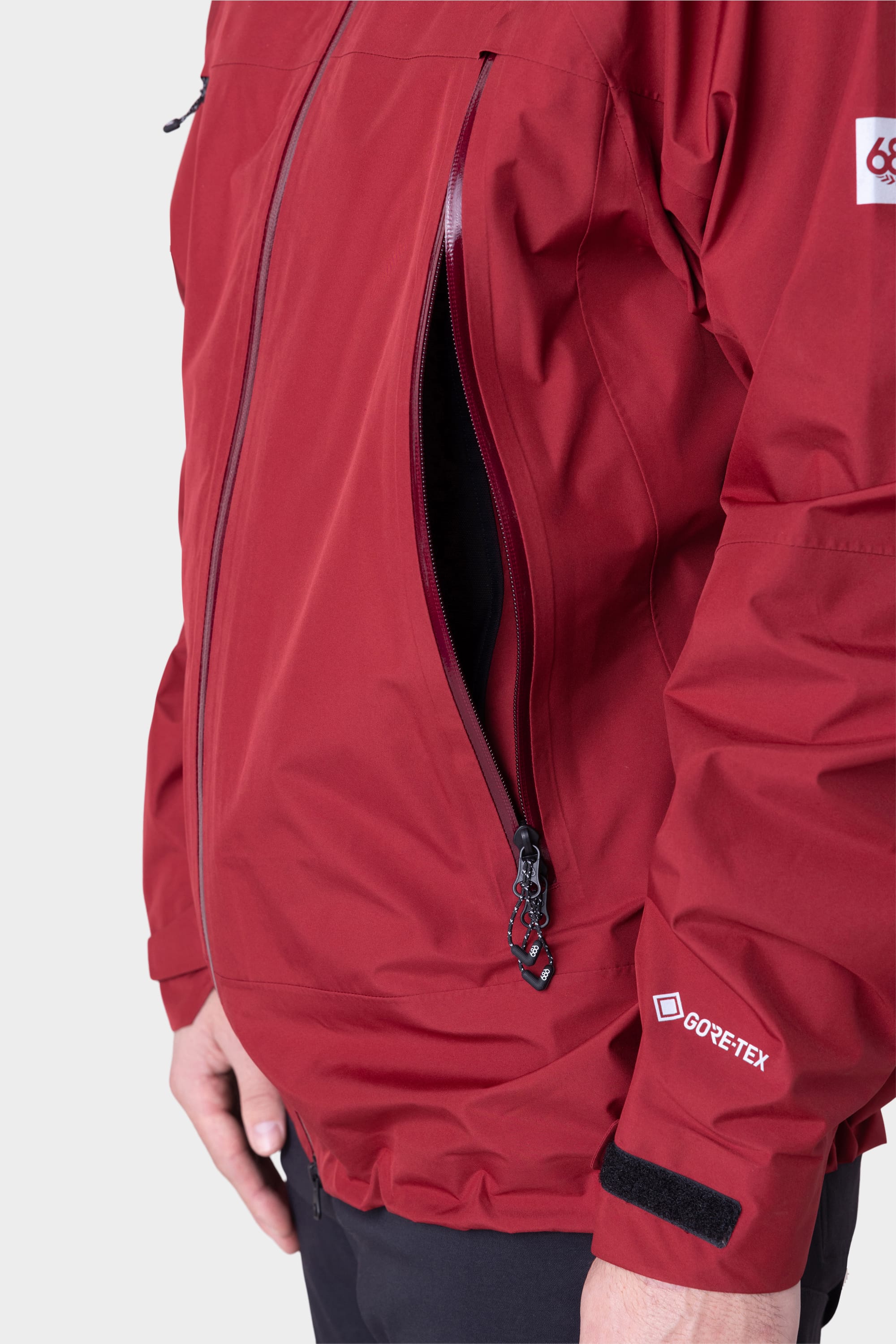 A person wears the 686 Mens GORE-TEX PACLITE® Jacket in red, with a partially unzipped side pocket showing its black inner lining. Only the torso and one hand are visible against a plain background. Brand: 686.