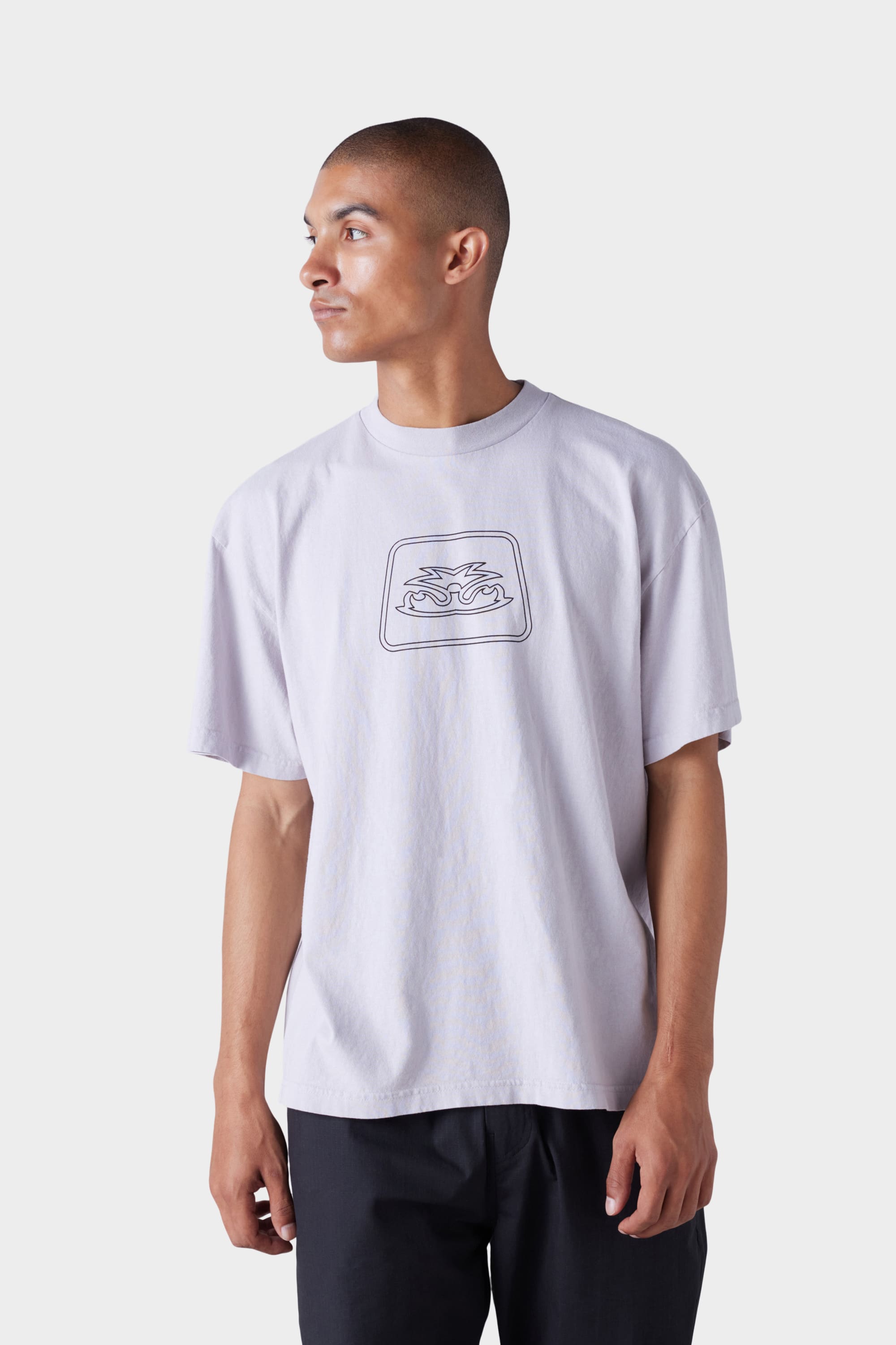 A man wearing the 686 Mens Jib Flower Outline Short Sleeve Tee by 686, a loose-fitting, light-colored T-shirt with a simple line art flower graphic on the chest, looks to his left against a plain light grey background.