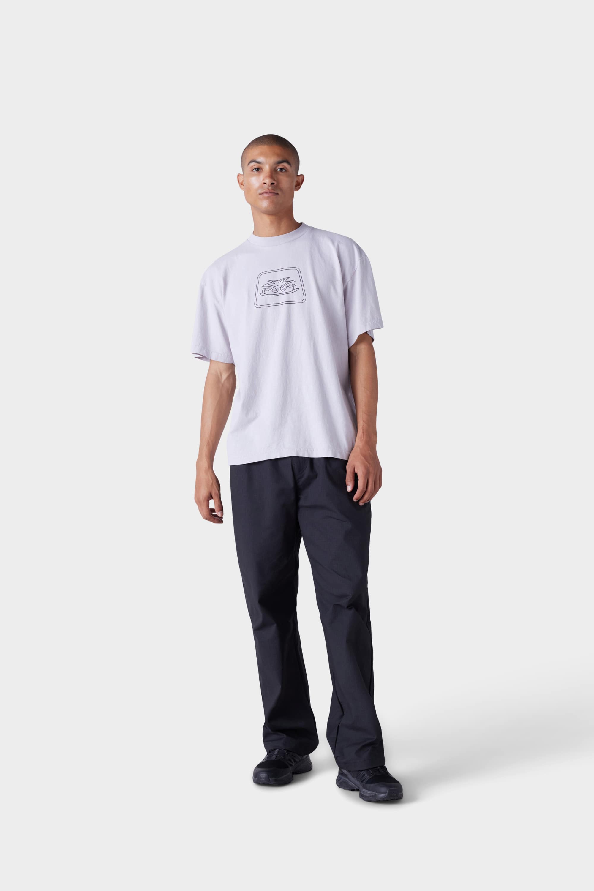 A person stands facing forward against a plain white background, wearing the 686 Mens Jib Flower Outline Short Sleeve Tee in light purple, loose black pants, and black sneakers.