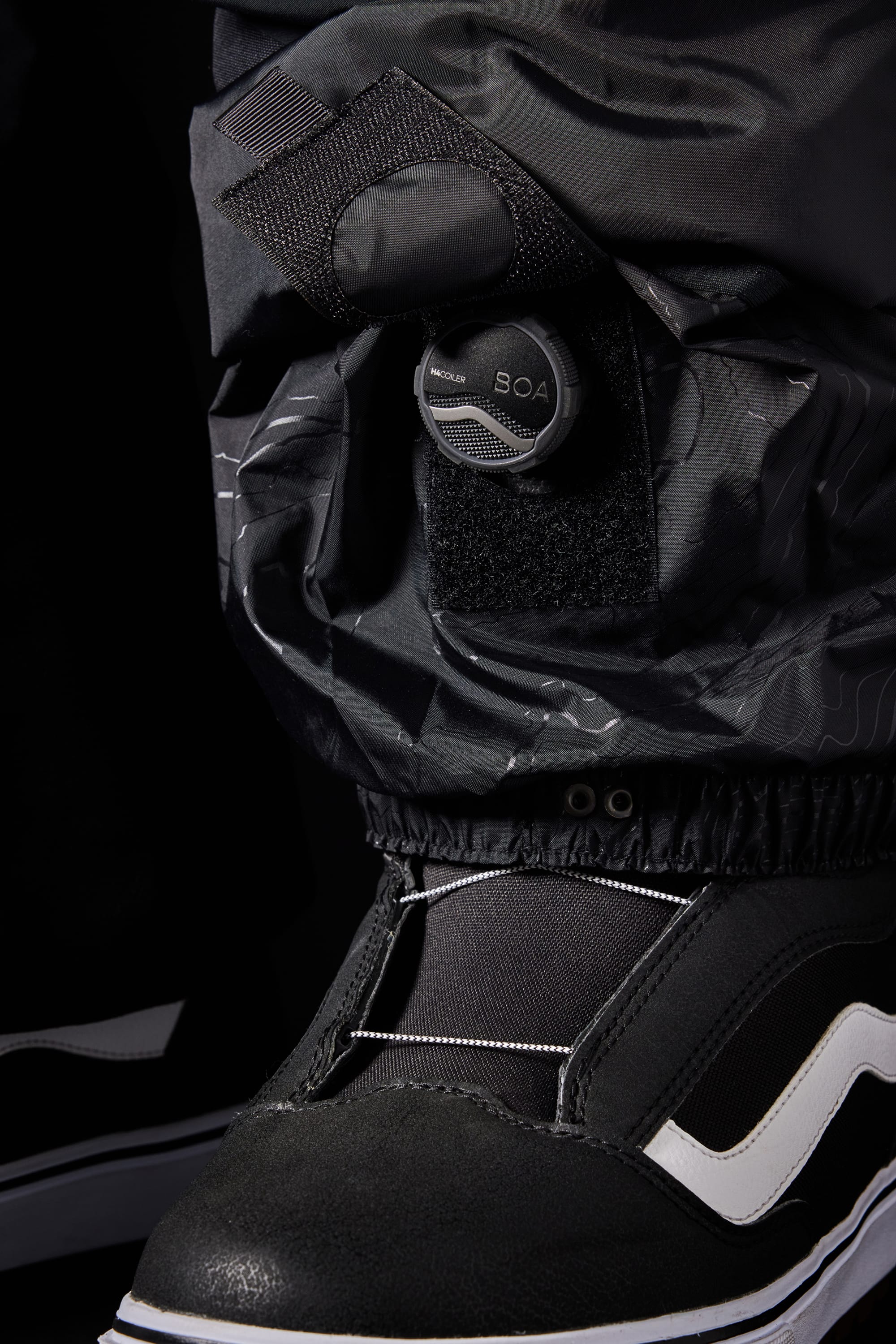 A close-up of a black snowboarding boot with a white stripe and Boa dial lacing system, with the 686 Men’s SMARTY® 3-in-1 Cargo Pant cuff resting above the boot.