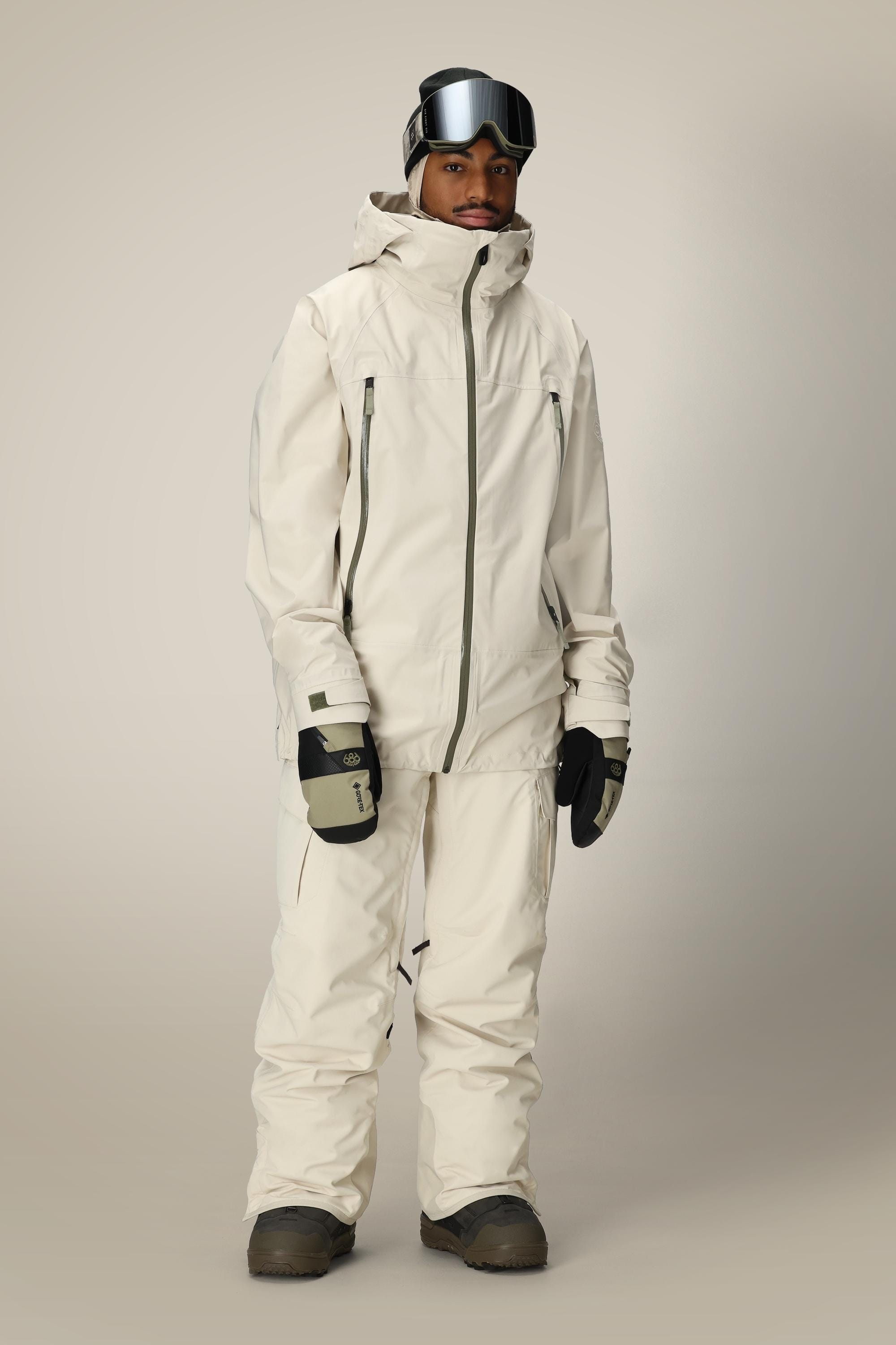 A person stands posing against a plain light background, dressed in the 686 Men’s SMARTY® 3-in-1 Cargo Pant, a cream-colored snowboarding jacket and pants, black gloves, boots, and black goggles resting on their forehead.