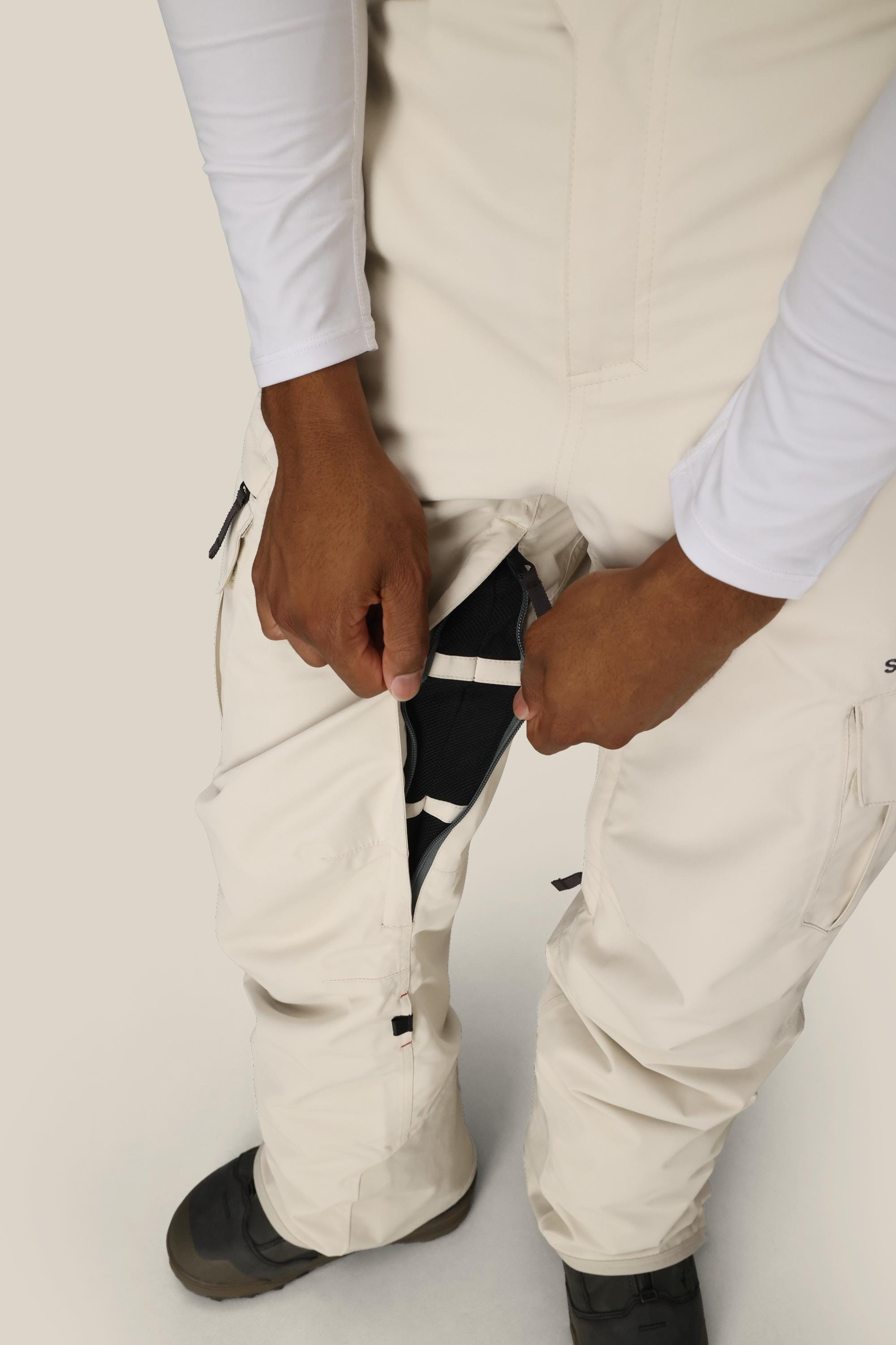 A person is unzipping a side vent on their white 686 Men’s SMARTY® 3-in-1 Cargo Pant, revealing black inner fabric. Only the lower torso, hands, and part of the legs are visible.