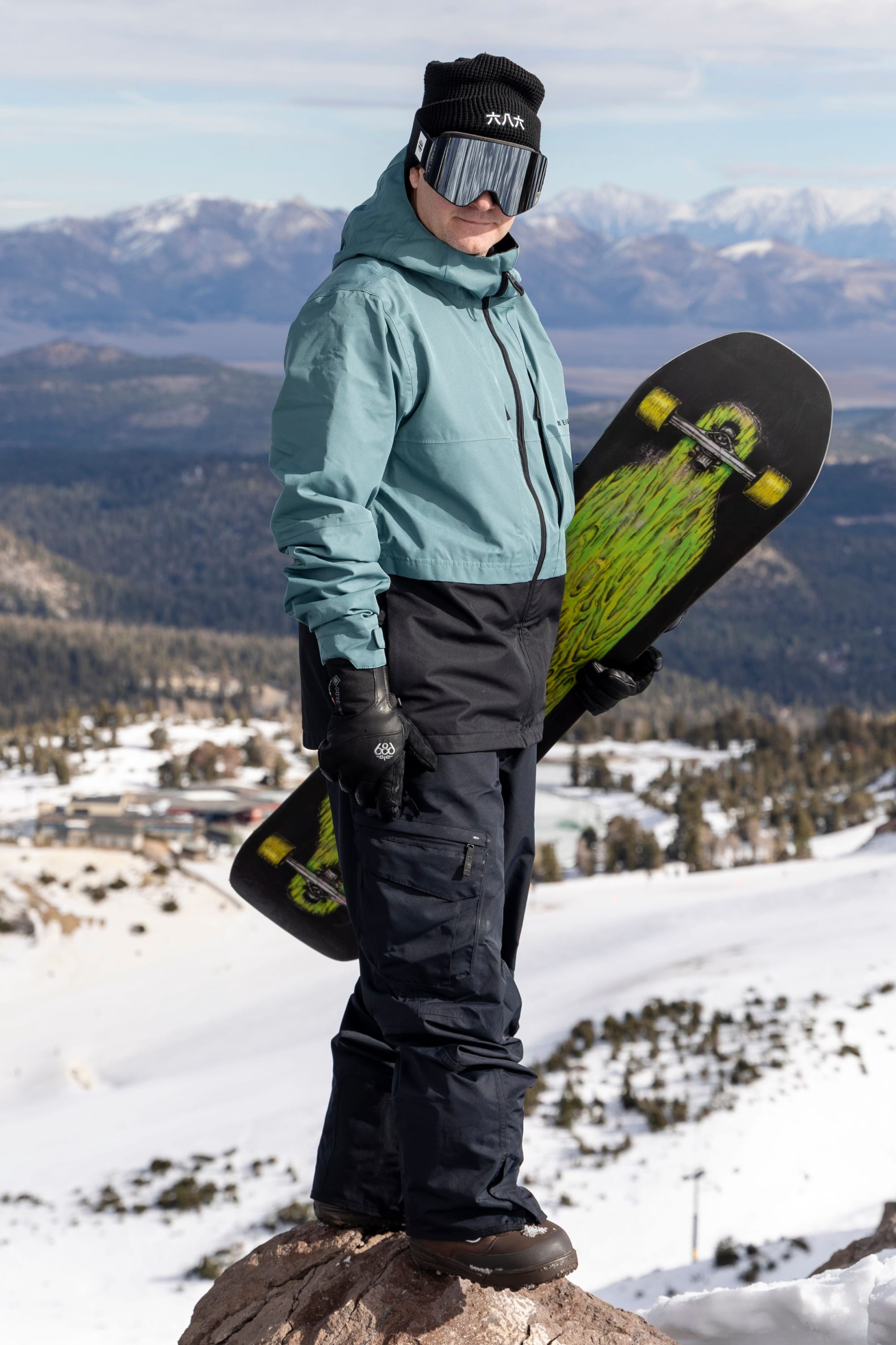 Wearing the 686 Men’s SMARTY® 3-in-1 Cargo Pant, a person in winter gear stands on a snowy mountain holding a green-accented snowboard, overlooking a breathtaking landscape of mountains and trees.