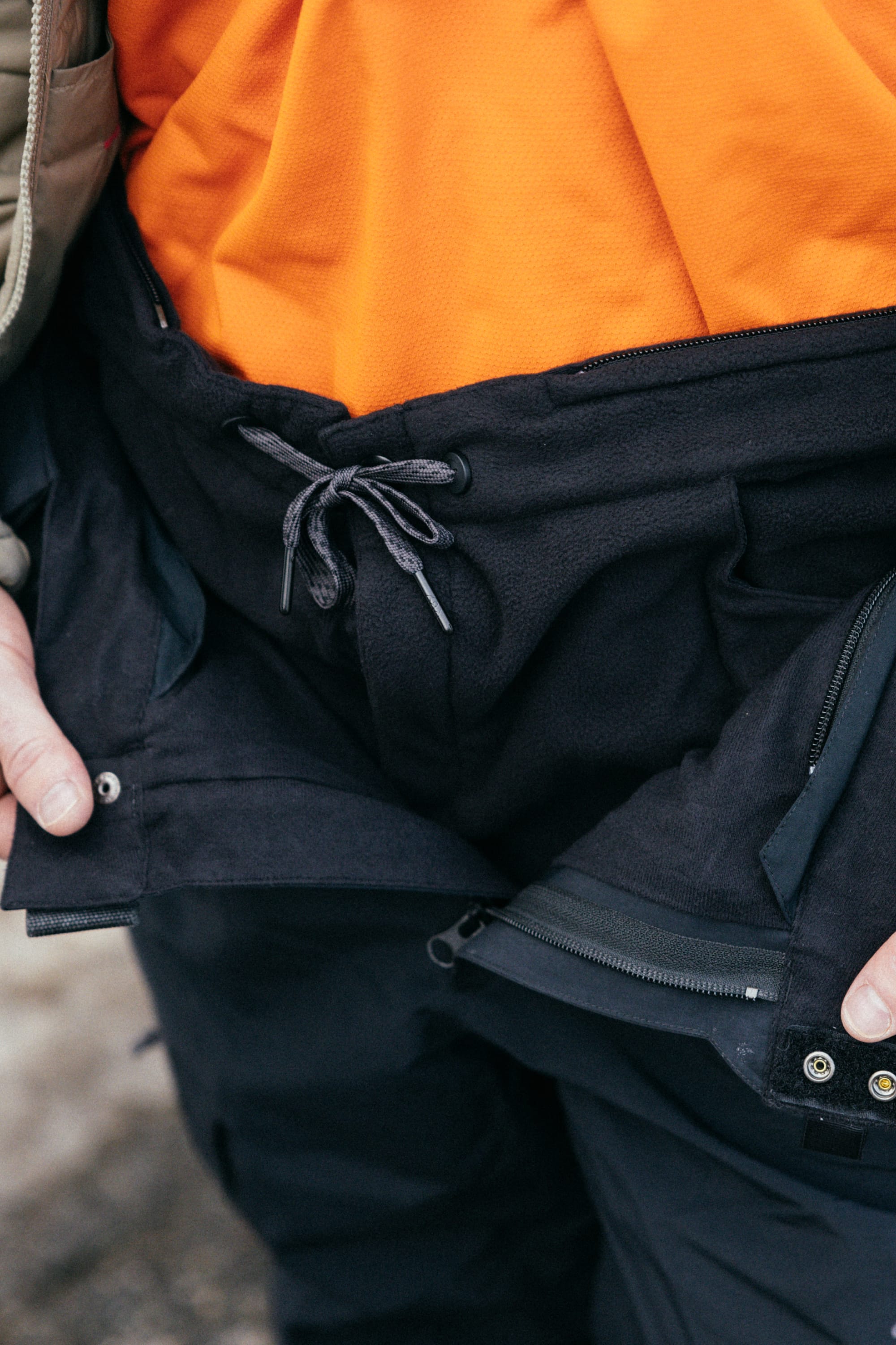 A close-up of a person wearing the 686 Men’s SMARTY® 3-in-1 Cargo Pant in black, paired with an orange shirt and a partially unzipped jacket, with their hands visible near the waistband.