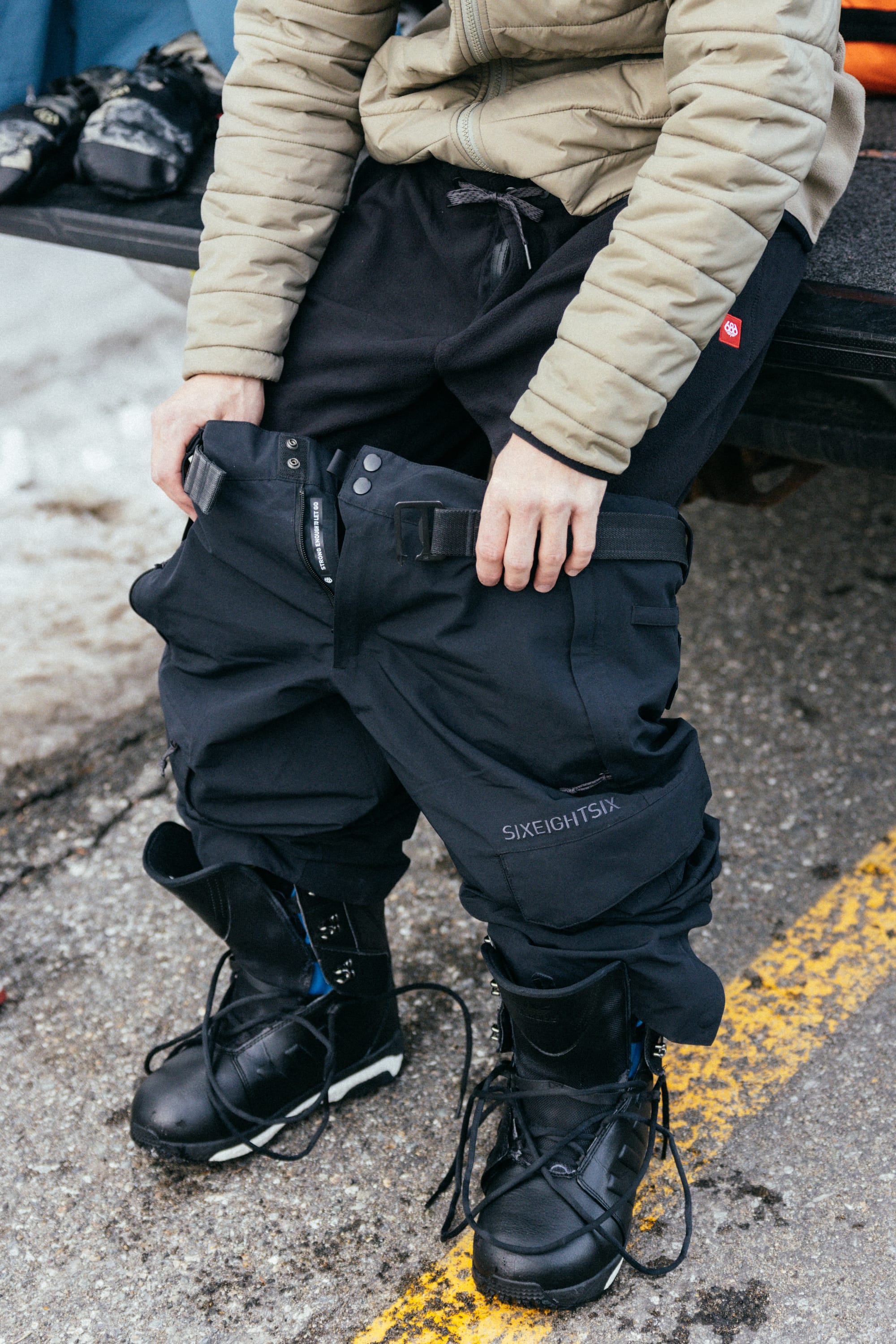 A person sits on a snowy roadside, pulling on black 686 Men’s SMARTY® 3-in-1 Cargo Pants over their black snow boots with untied laces. They are wearing a beige jacket, and the word “686” is visible on the pants.