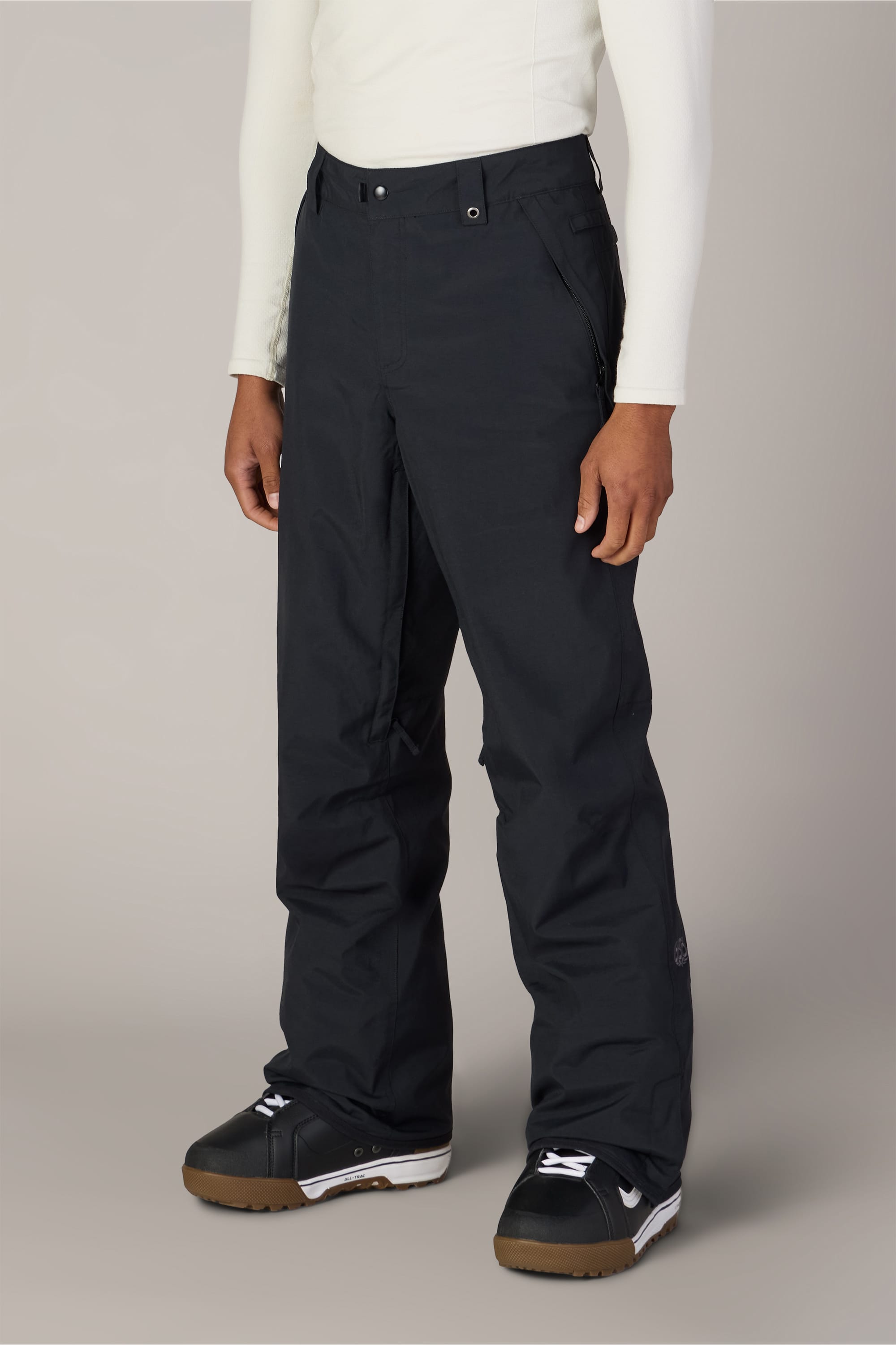 A person wearing the 686 Men’s Standard Shell Pant in black, a white long-sleeve shirt, and black snow boots stands against a plain light gray background.