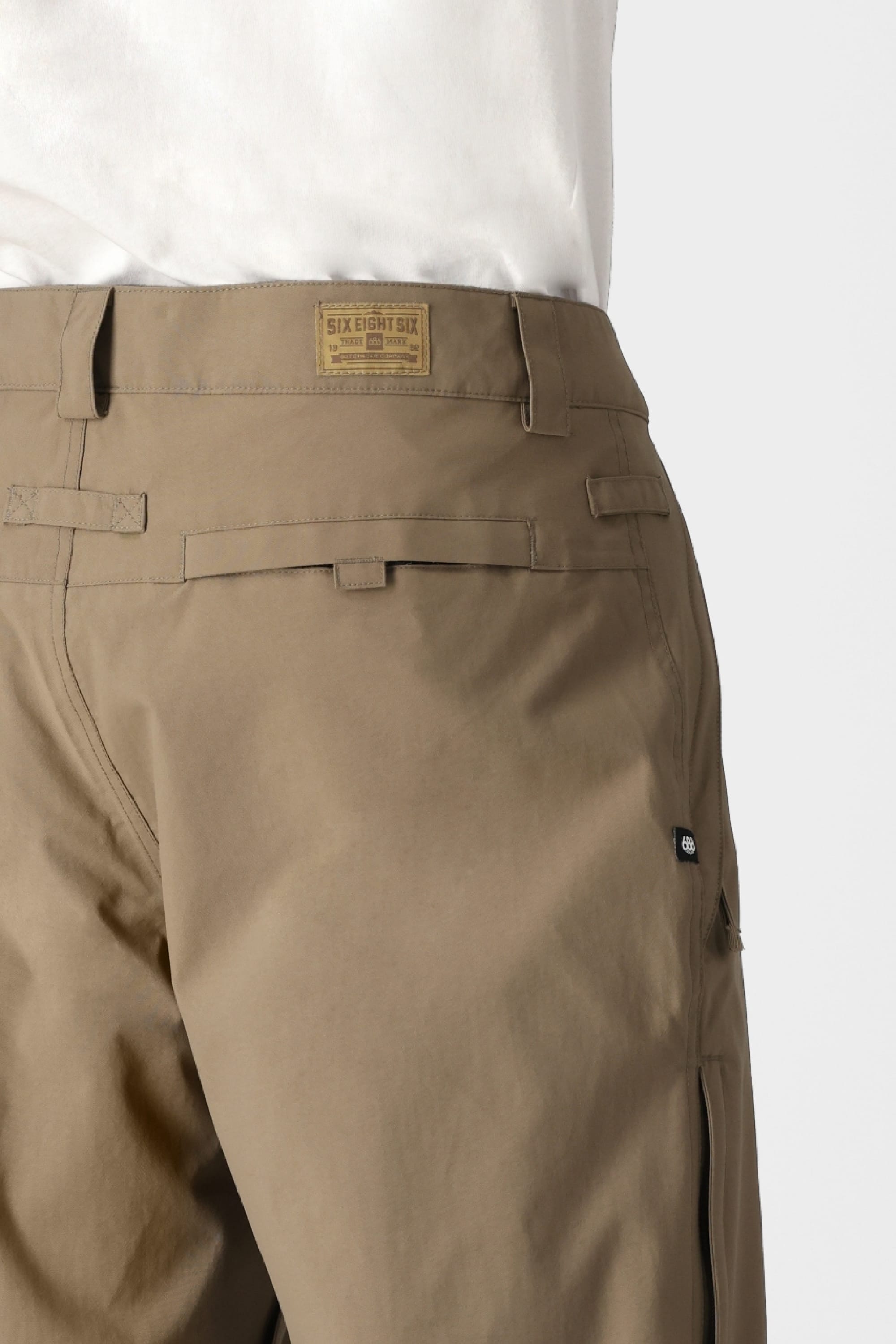 A person wearing the 686 Men’s Standard Shell Pant in light brown, featuring a visible 686 label on the waistband, stands against a plain light gray background.