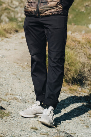 A person stands on a rocky outdoor path, wearing 686 Men's Everywhere® Pant - Slim Fit in black, light gray sneakers, and a brown puffer jacket. The image is cropped to show only the person’s lower body and legs.