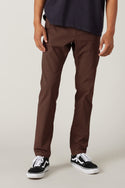 A person in 686 Men's Everywhere® Pant - Slim Fit (brown) by 686, with a dark shirt and black-and-white sneakers, stands against a plain light background. Their hands are at their sides and their face is not visible.