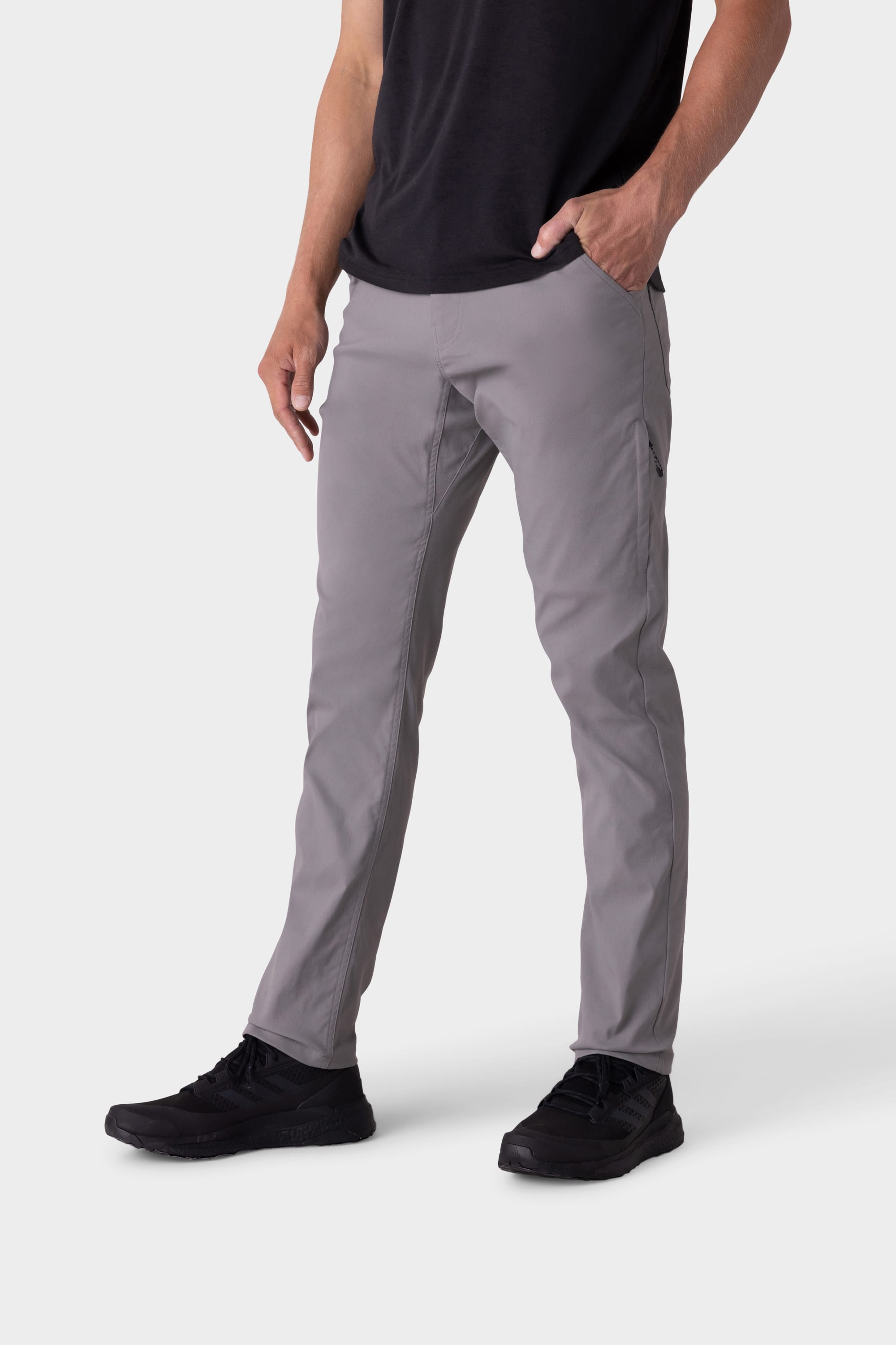 A person wears 686 Mens Everywhere Pant - Slim Fit in light gray, a black t-shirt, and black sneakers, standing with one hand in their pocket against a plain white background.