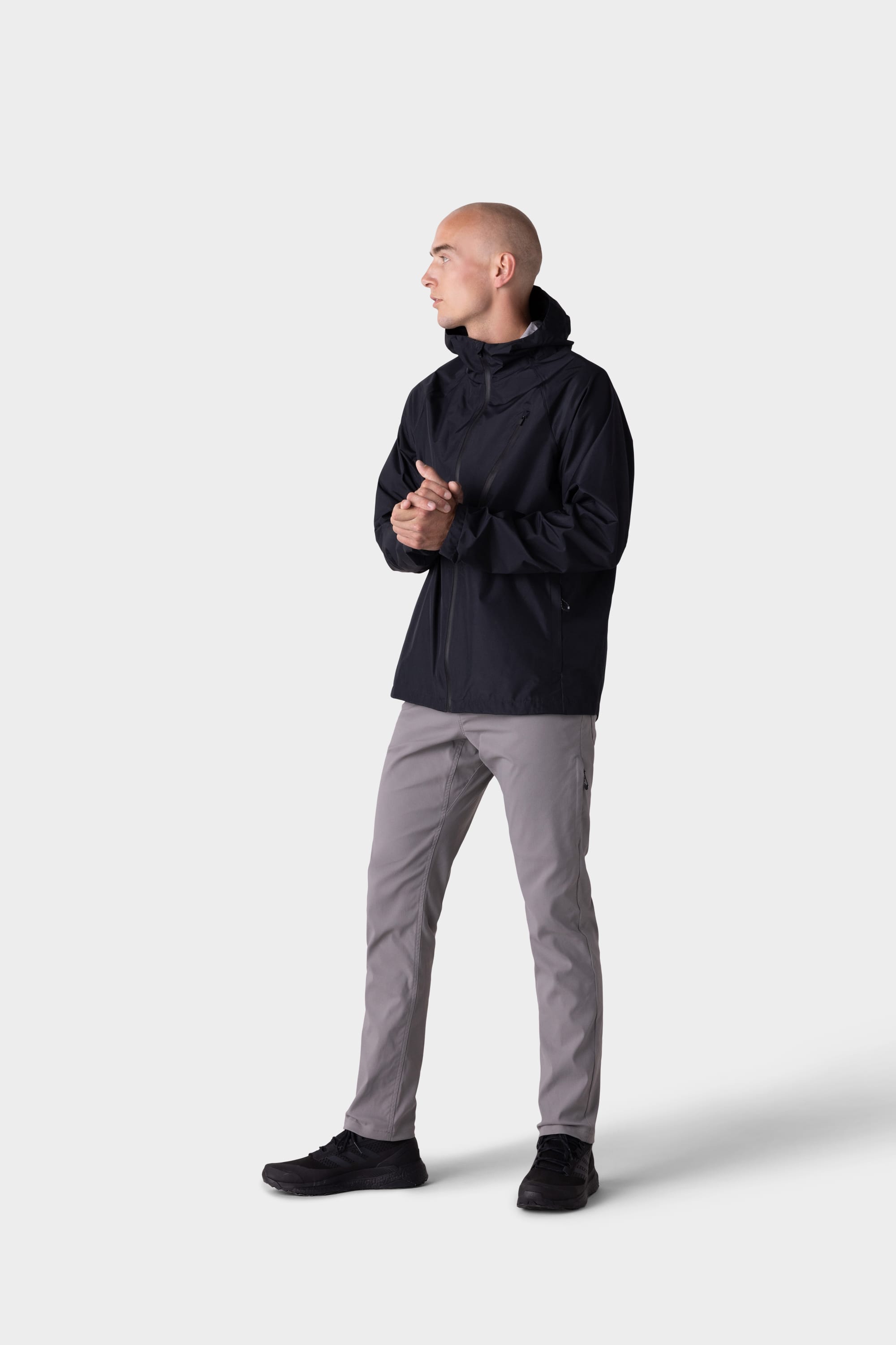 A man in a Slim Fit black hooded jacket, 686 Mens Everywhere Pant - Slim Fit by 686 in light gray, and black shoes stands against a plain light background, looking to his left with his hands clasped.
