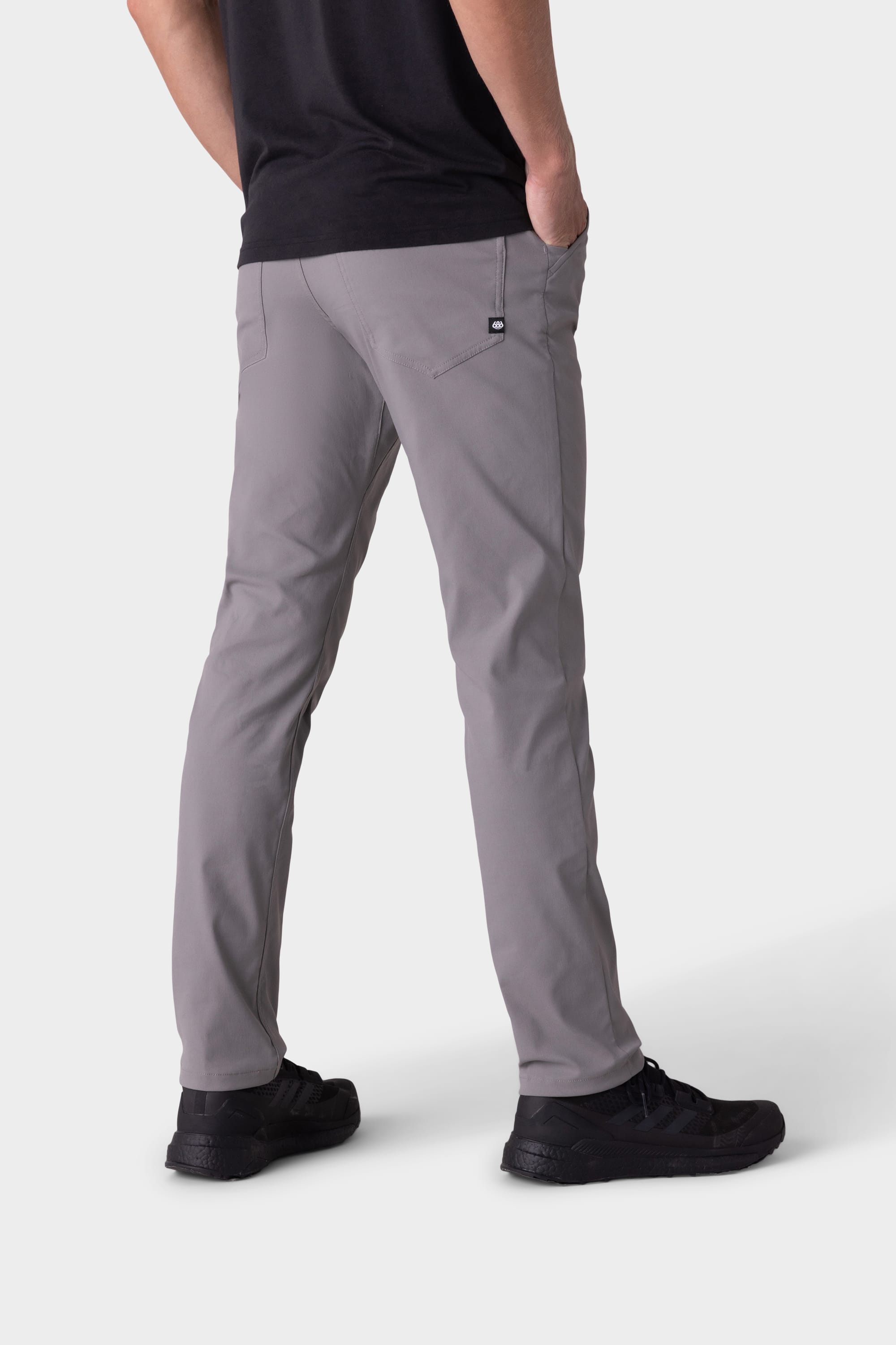 A person stands with hands in pockets, wearing 686 Mens Everywhere Pant - Slim Fit in light gray, paired with a black shirt and black shoes, shown from the back against a plain white background.