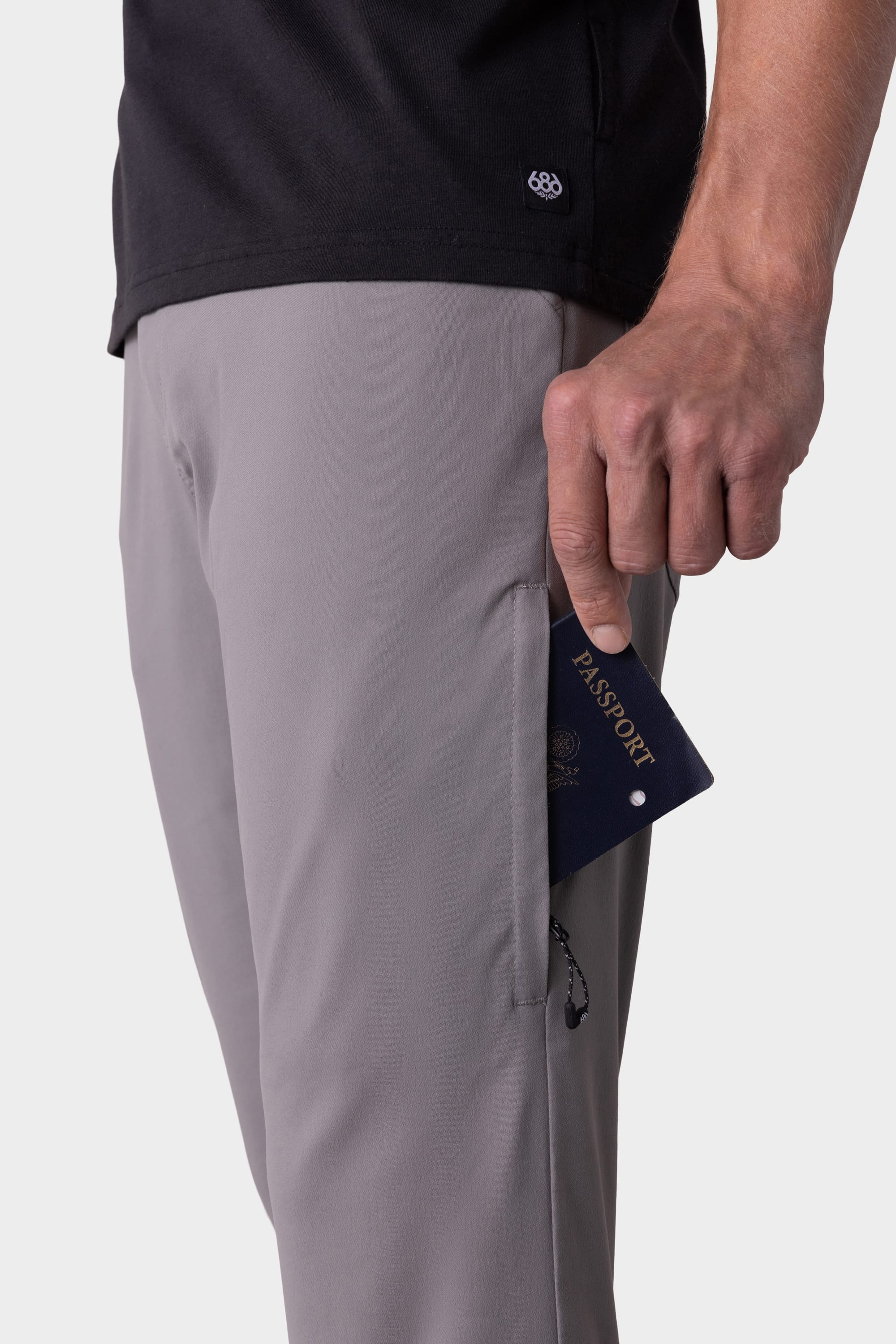 A person in stain-resistant gray 686 Mens Everywhere Pant - Slim Fit and a black shirt holds a passport partially tucked into the pant’s front pocket.